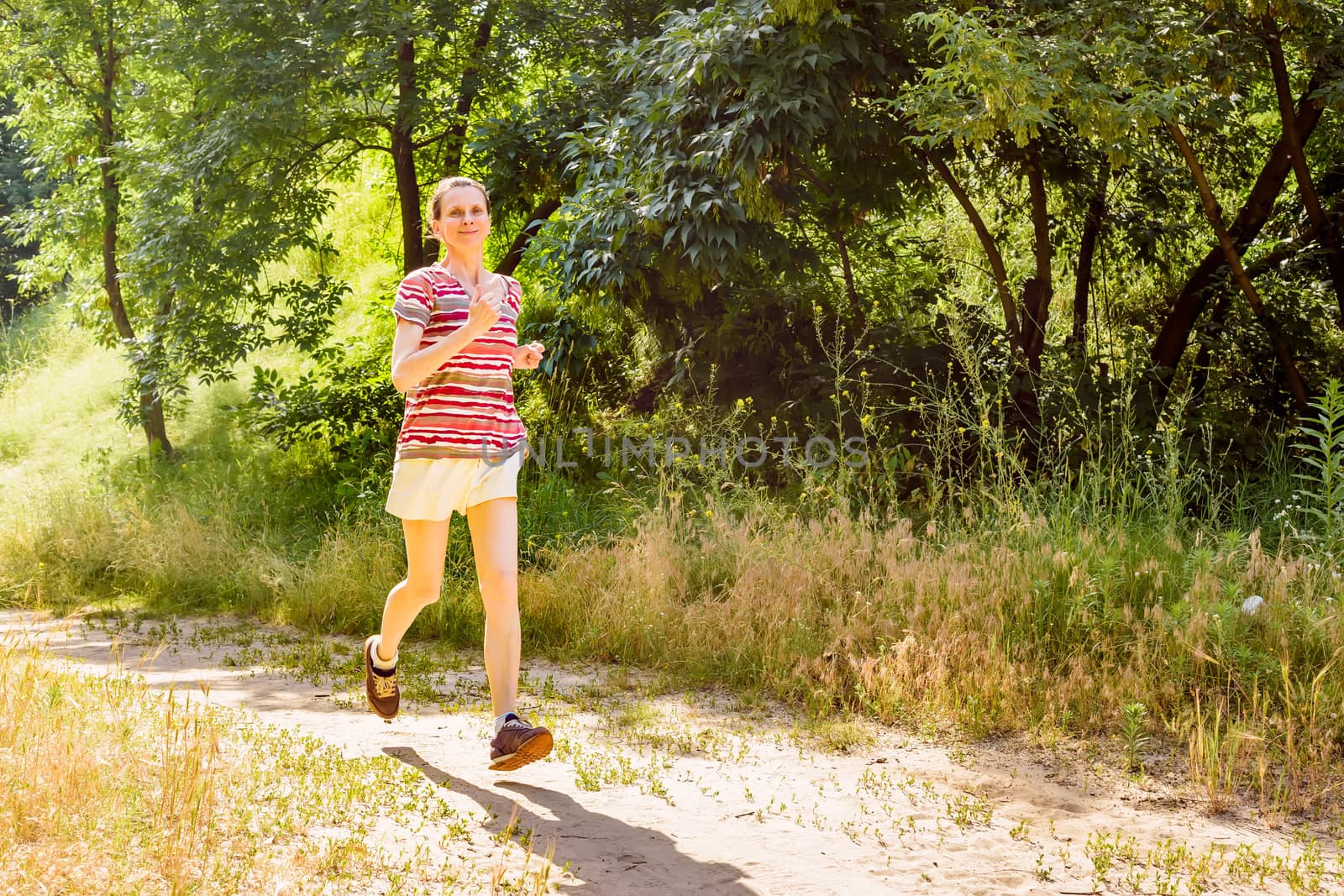 A happy senior woman is running with the sun in backlight, toward the shadow, in the forest during a warm summer day