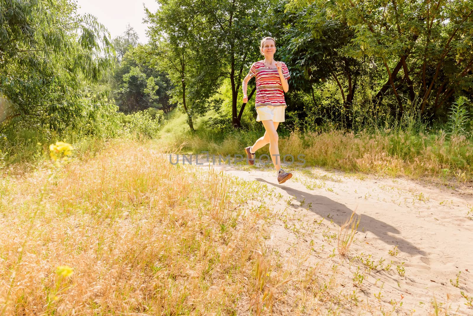 A happy senior woman is running with the sun in backlight, toward the shadow, in the forest during a warm summer day