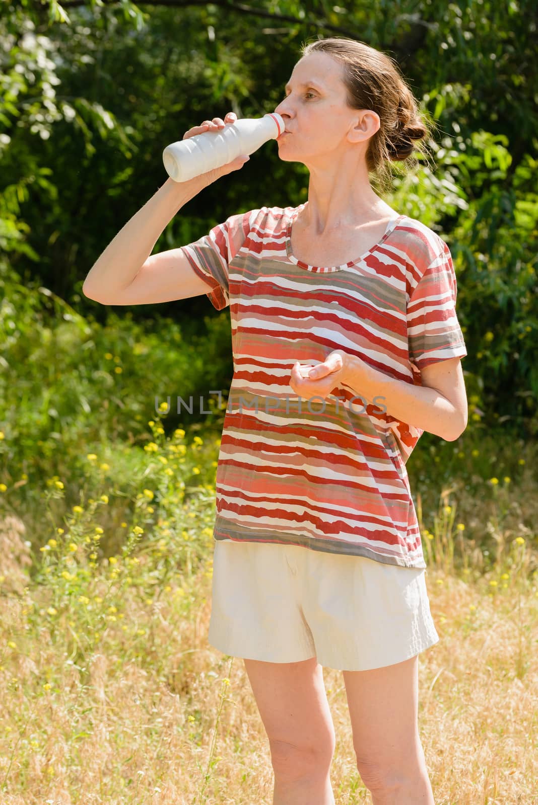A senior woman is drinking water or yogurth to quench her thirst after a long run in the forest