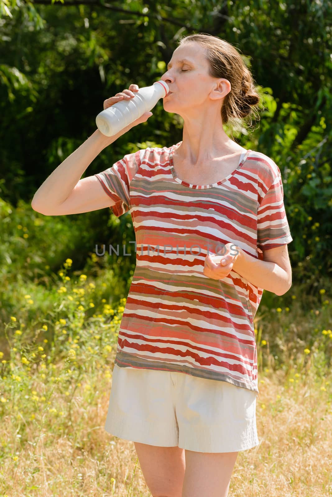 A senior woman is drinking water or yogurth to quench her thirst after a long run in the forest