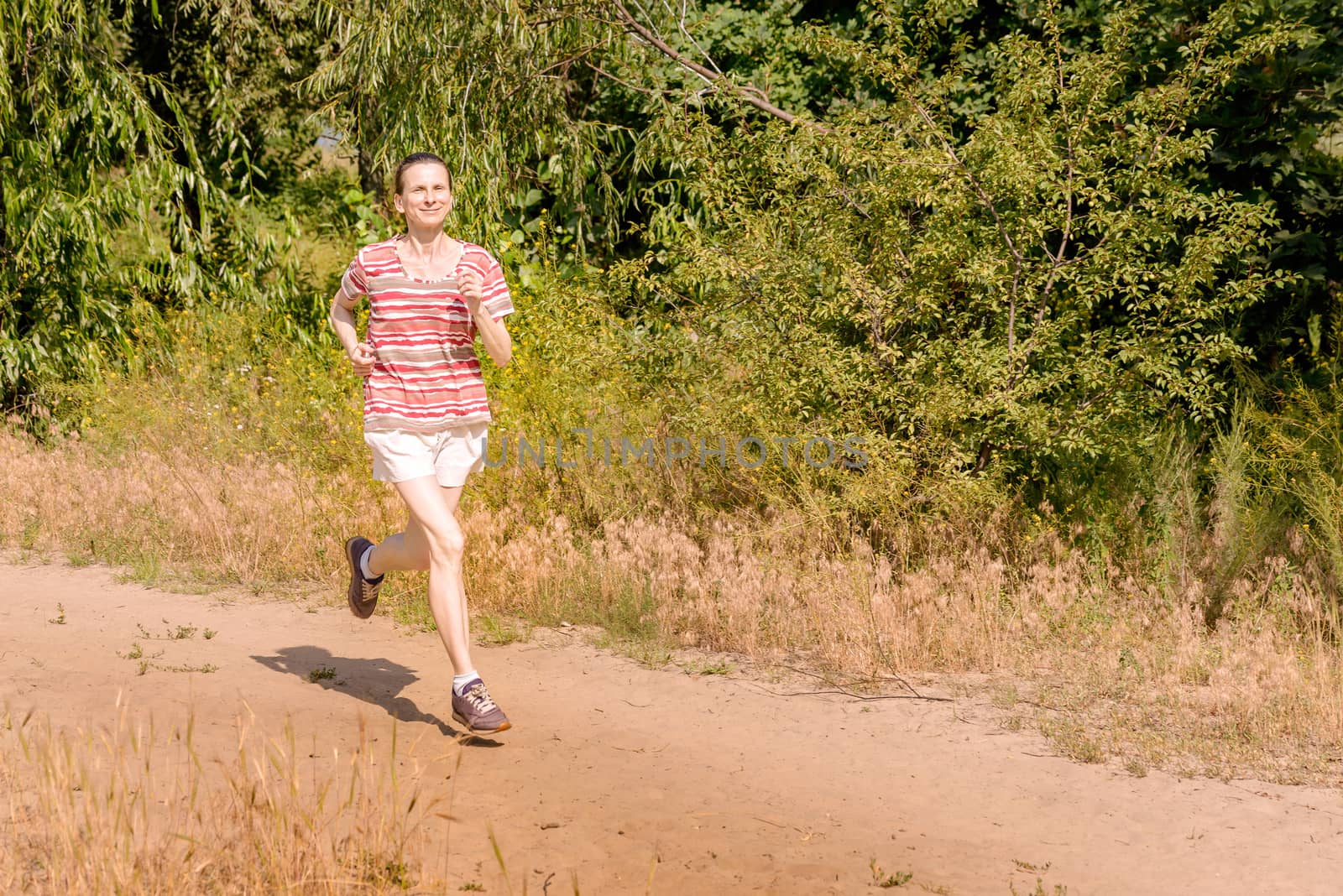 A happy senior woman is running under the sun in the forest during a warm summer day