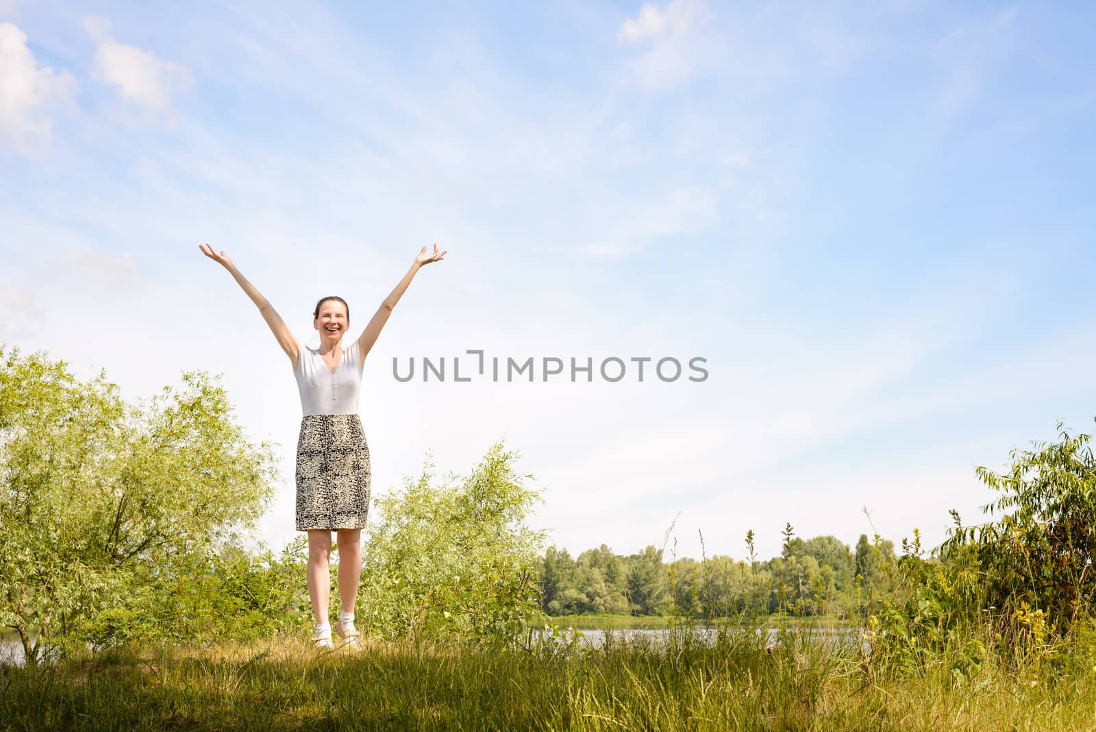 A happy adult woman standing close to the Dnieper river in Kiev, Ukraine, with the arms raised up forming the letter V, under a soft cloudy summer sky