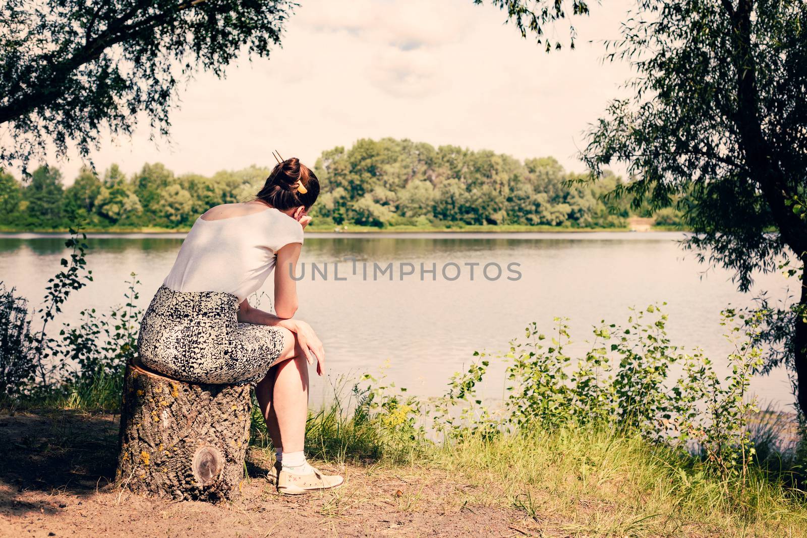 A sad and nostalgic woman is sitting close to the Dnieper river in Kiev, Ukraine. She is thinking while watching or observing far in the distance, under a warm and soft summer sun