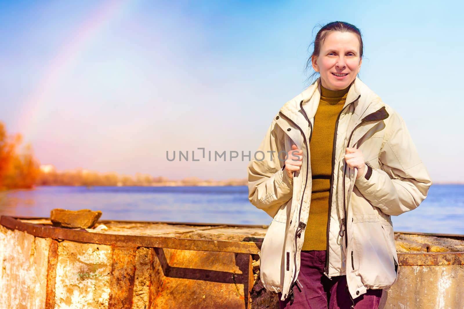 A natural mature woman smiling near the river. There is a light rainbow in the clear sky