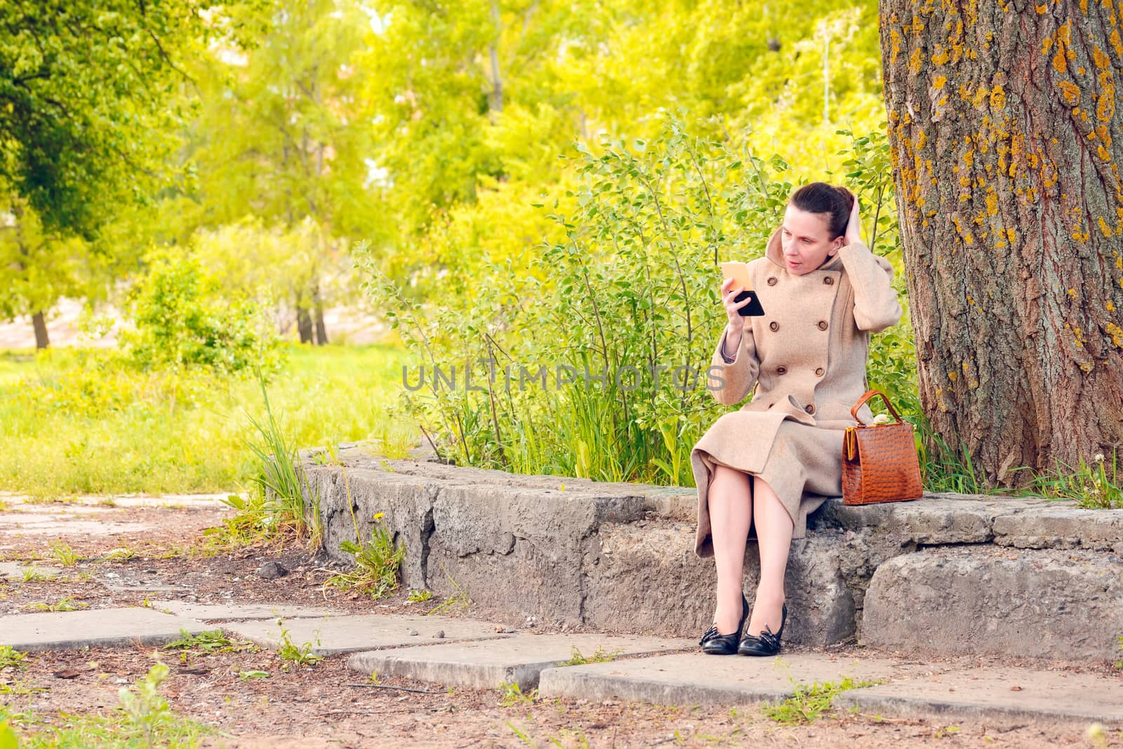 Elegant businesswoman sitting on a stone wall during a sunny spring day, and setting her hair looking herself in a little mirror