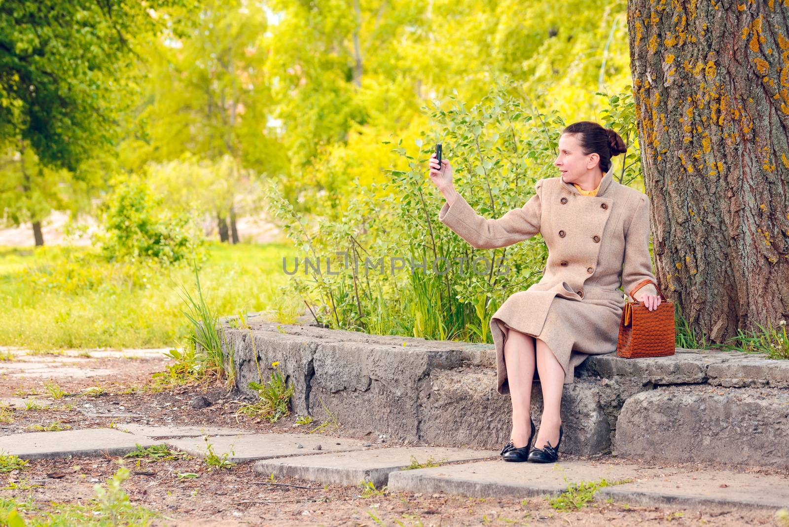 Elegant businesswoman sitting on a stone wall during a sunny spring day, and shooting a photo with her mobile phone