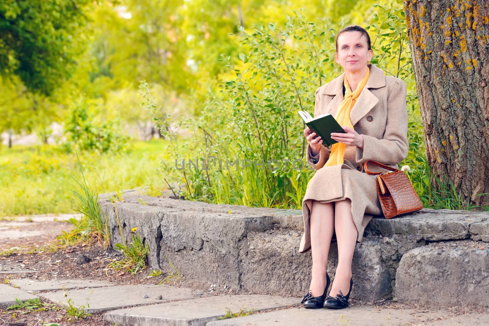 Elegant businesswoman sitting on a stone wall with a book during a sunny spring day, and watching at the sky