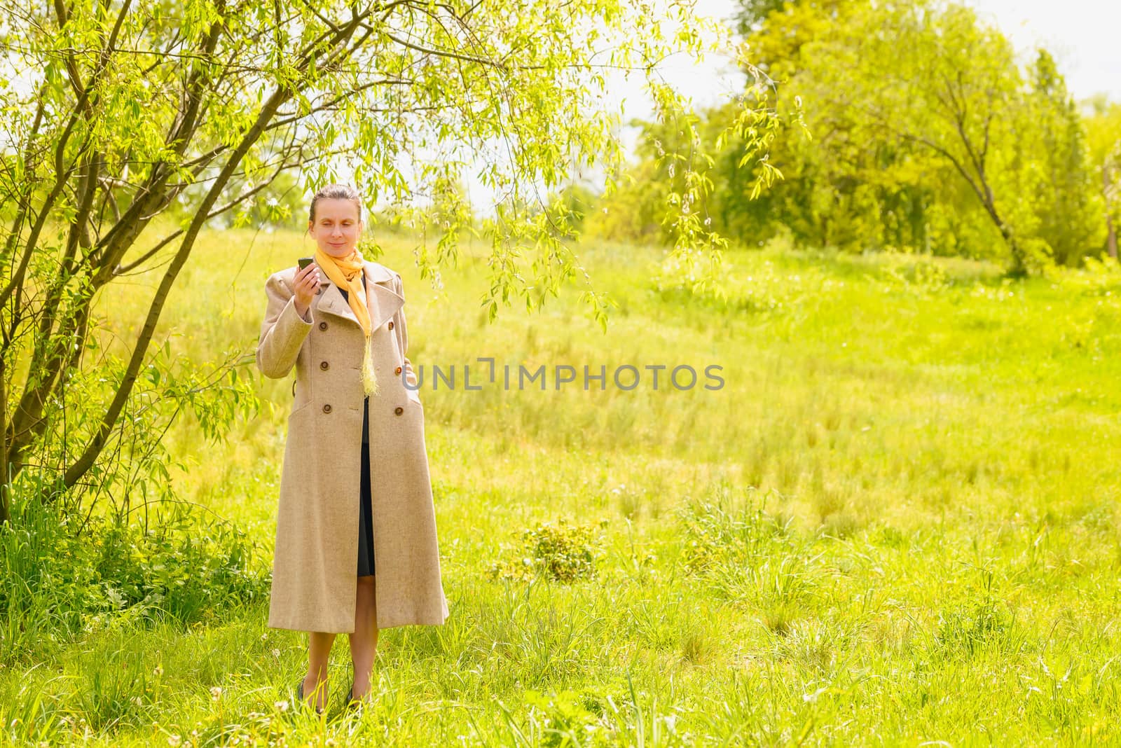An elegant senior businesswoman, with a coat and a yellow scarf, looking at her mobile phone in the park under the spring sun
