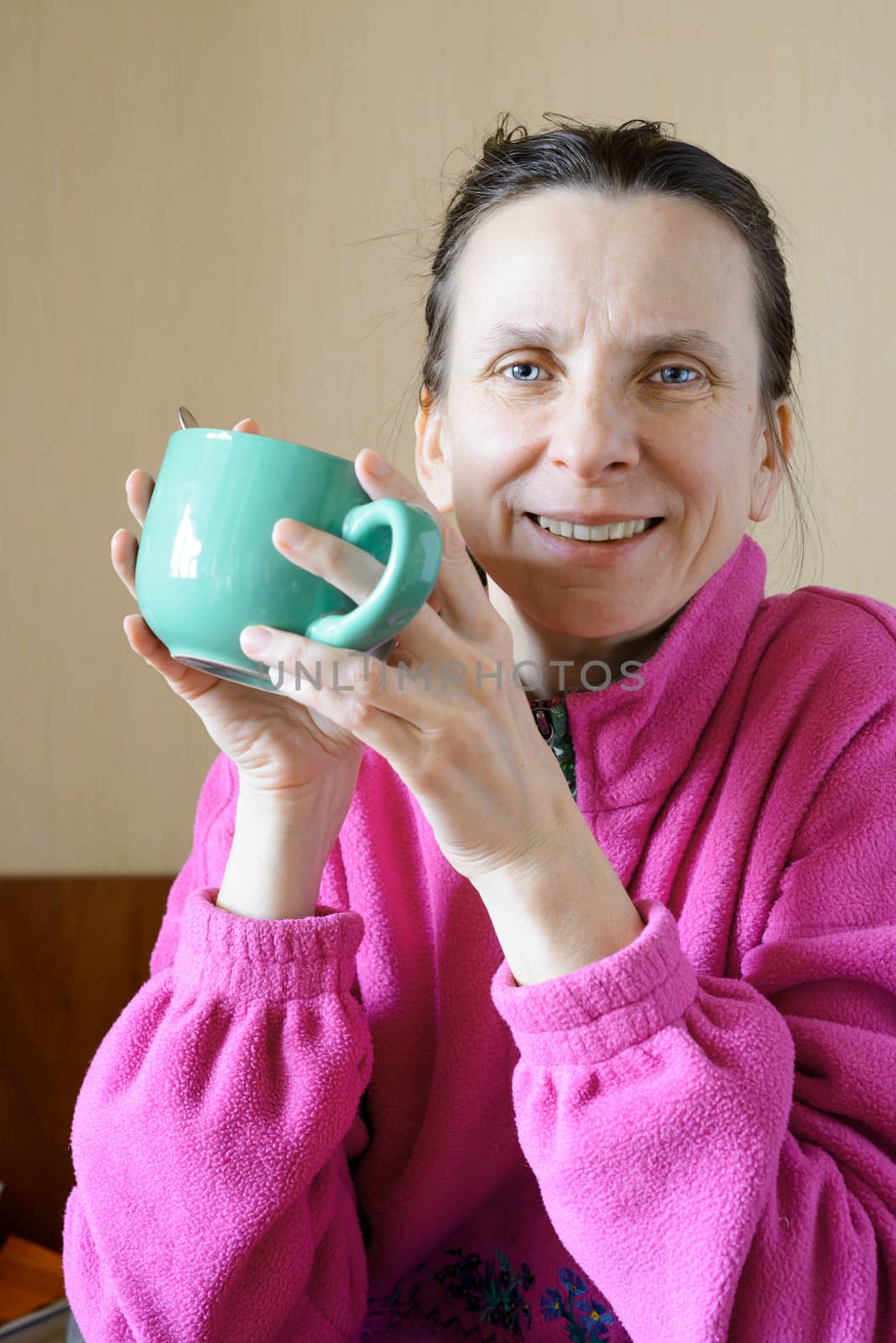 A smiling mature woman wearing a pink pull-over and drinking coffee in a green cup at morning for breakfast