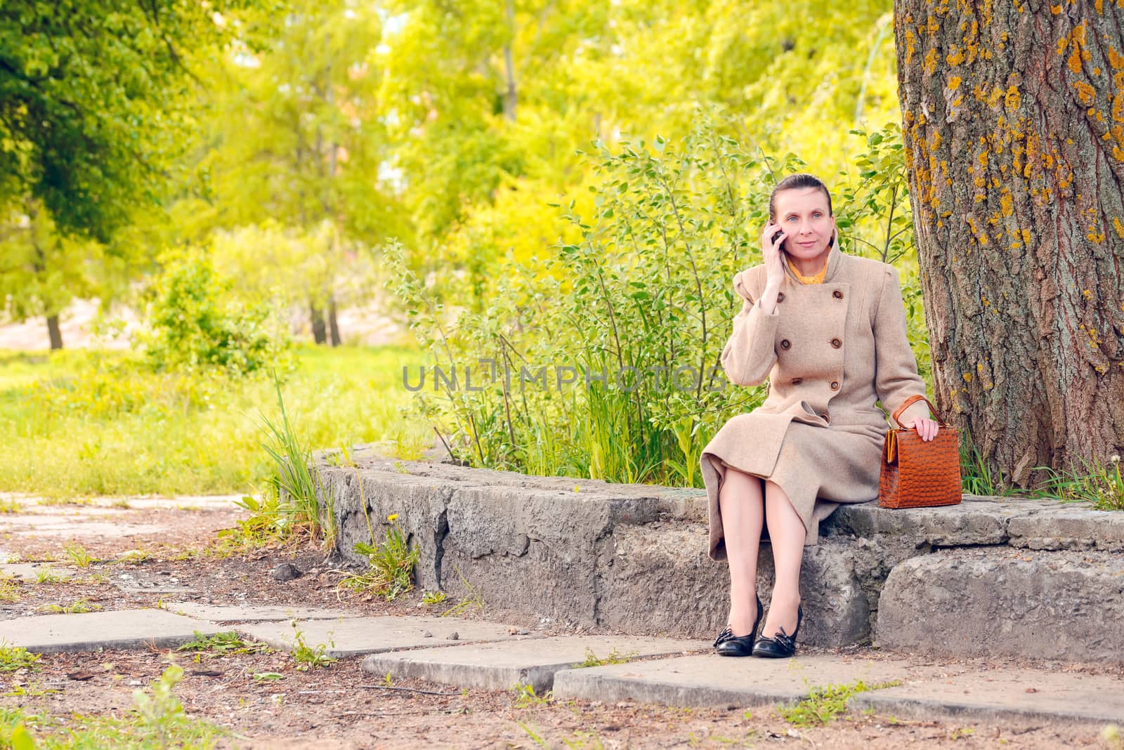 Elegant businesswoman sitting on a stone wall during a sunny spring day, and phoning to a customer