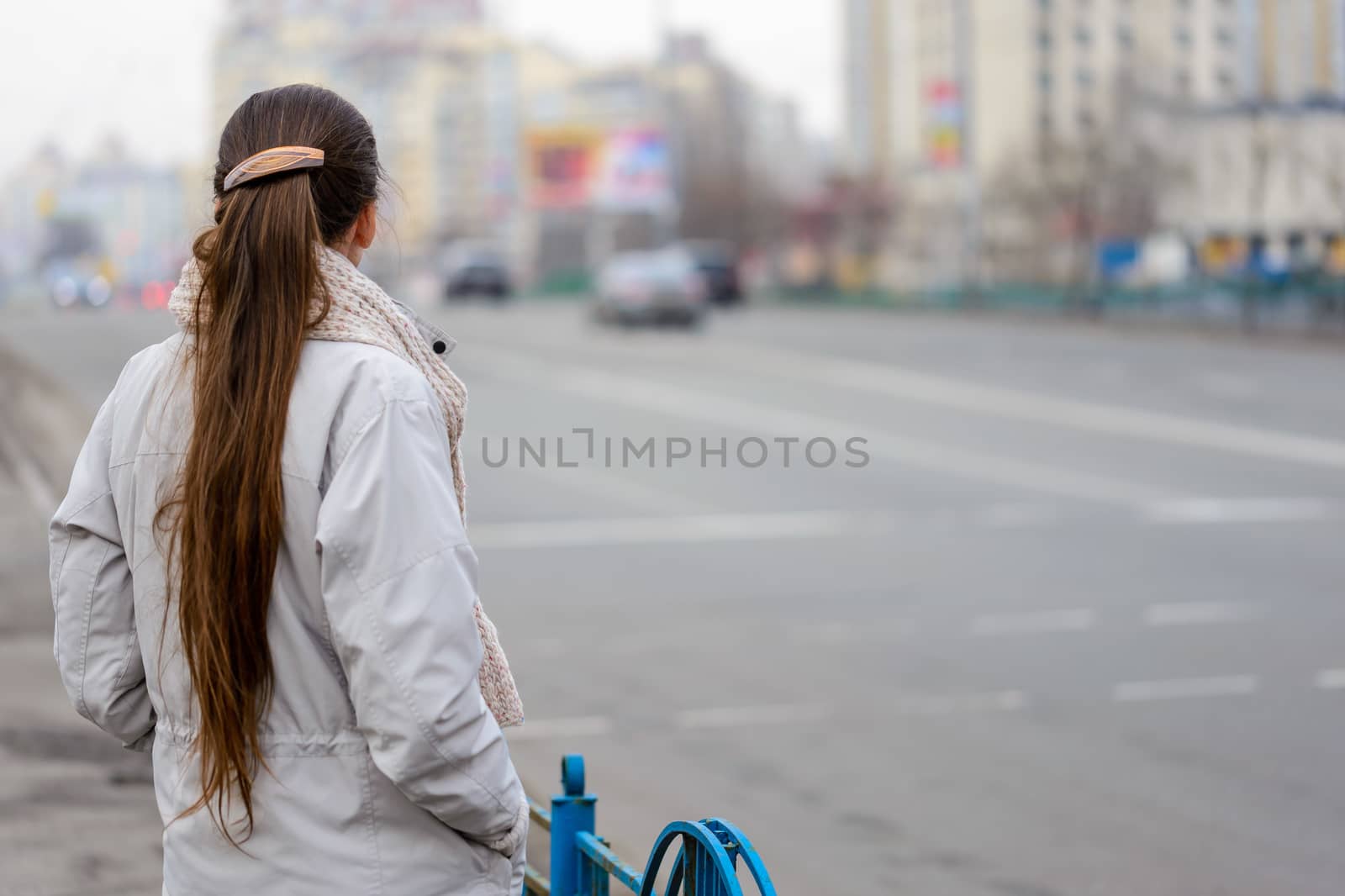 A woman with a ponytail stands, waiting or watching, close to the street with cars in the city