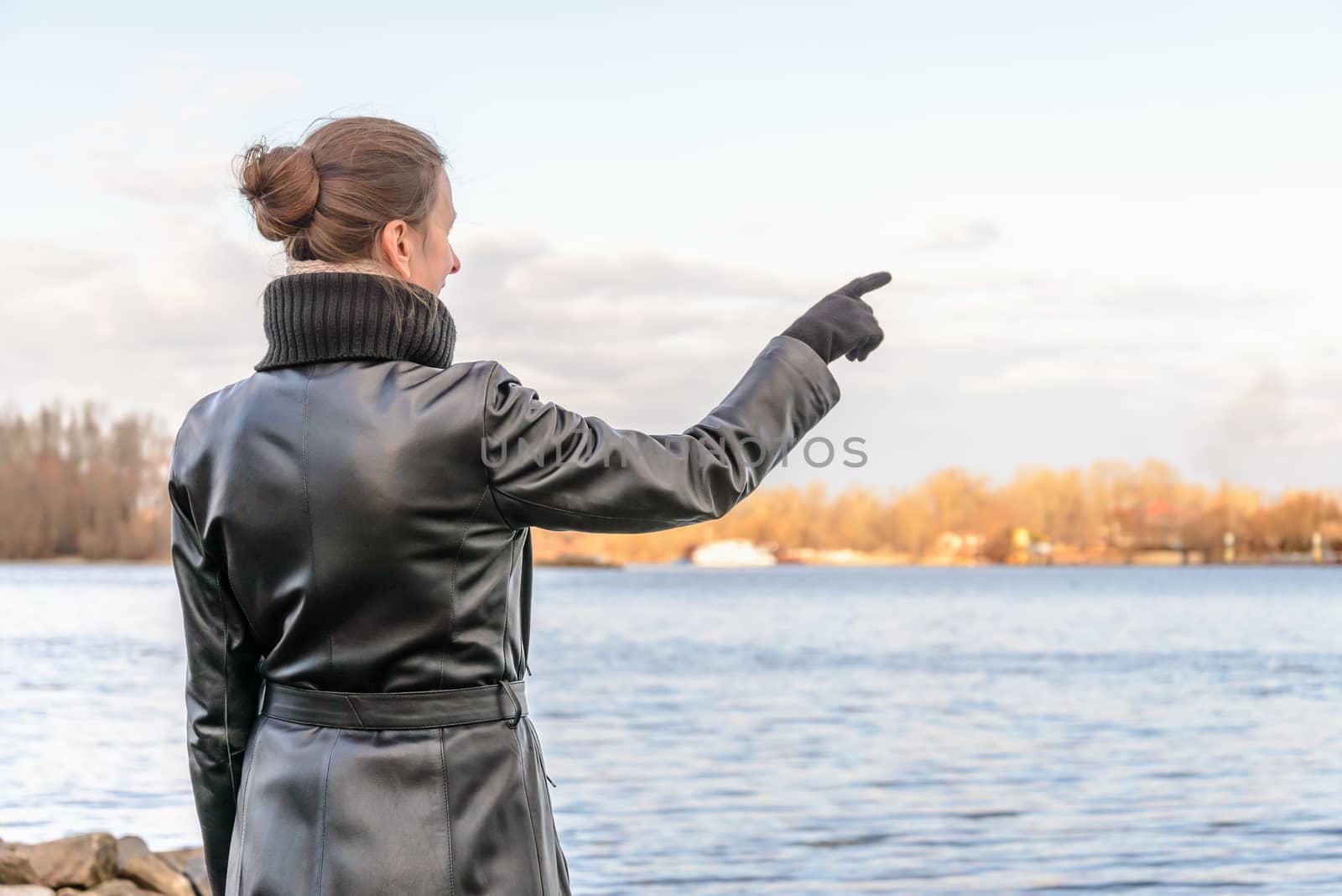 An adult with a chignon and wearing a black leather coat stays close to the river and point her finger to indicate something interesting on the opposite bank