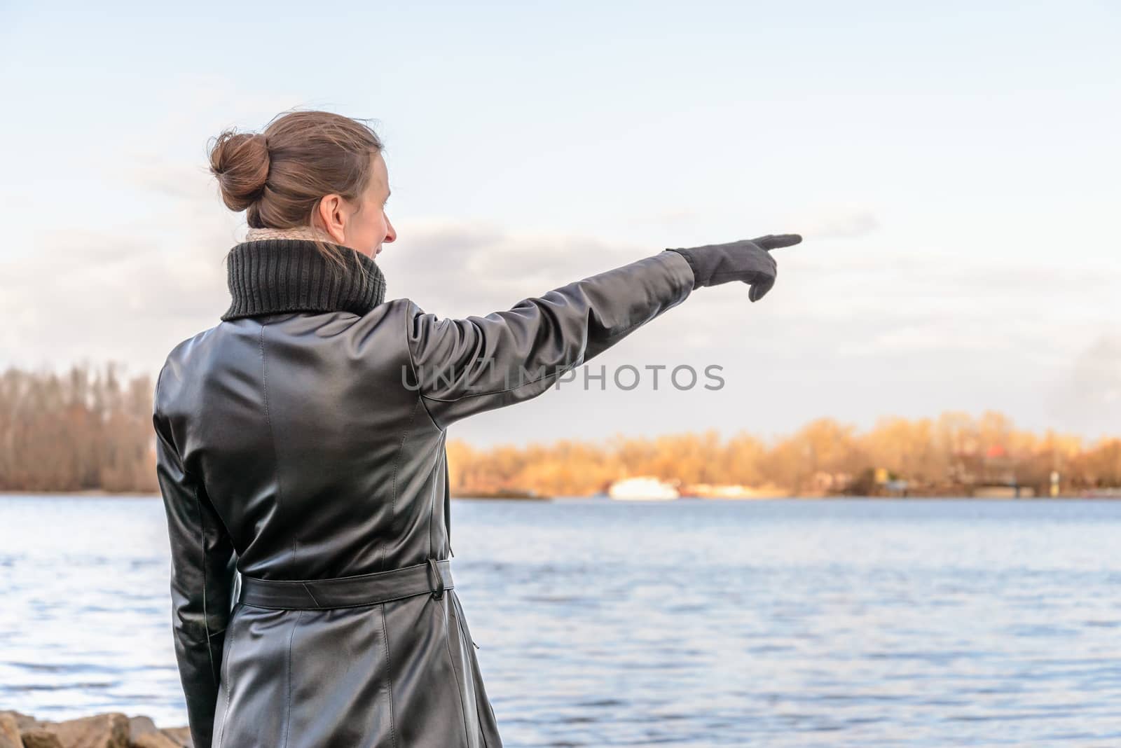 An adult with a chignon and wearing a black leather coat stays close to the river and point her finger to indicate something interesting on the opposite bank