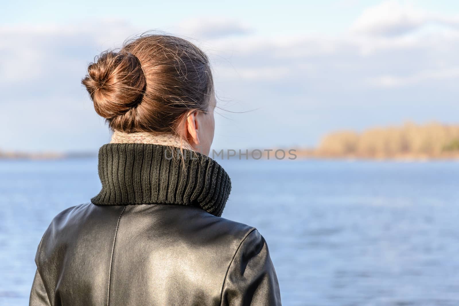 A woman with a chignon and a black leather coat is watching the landscape close to the lake or the river during a sunny day with big white clouds