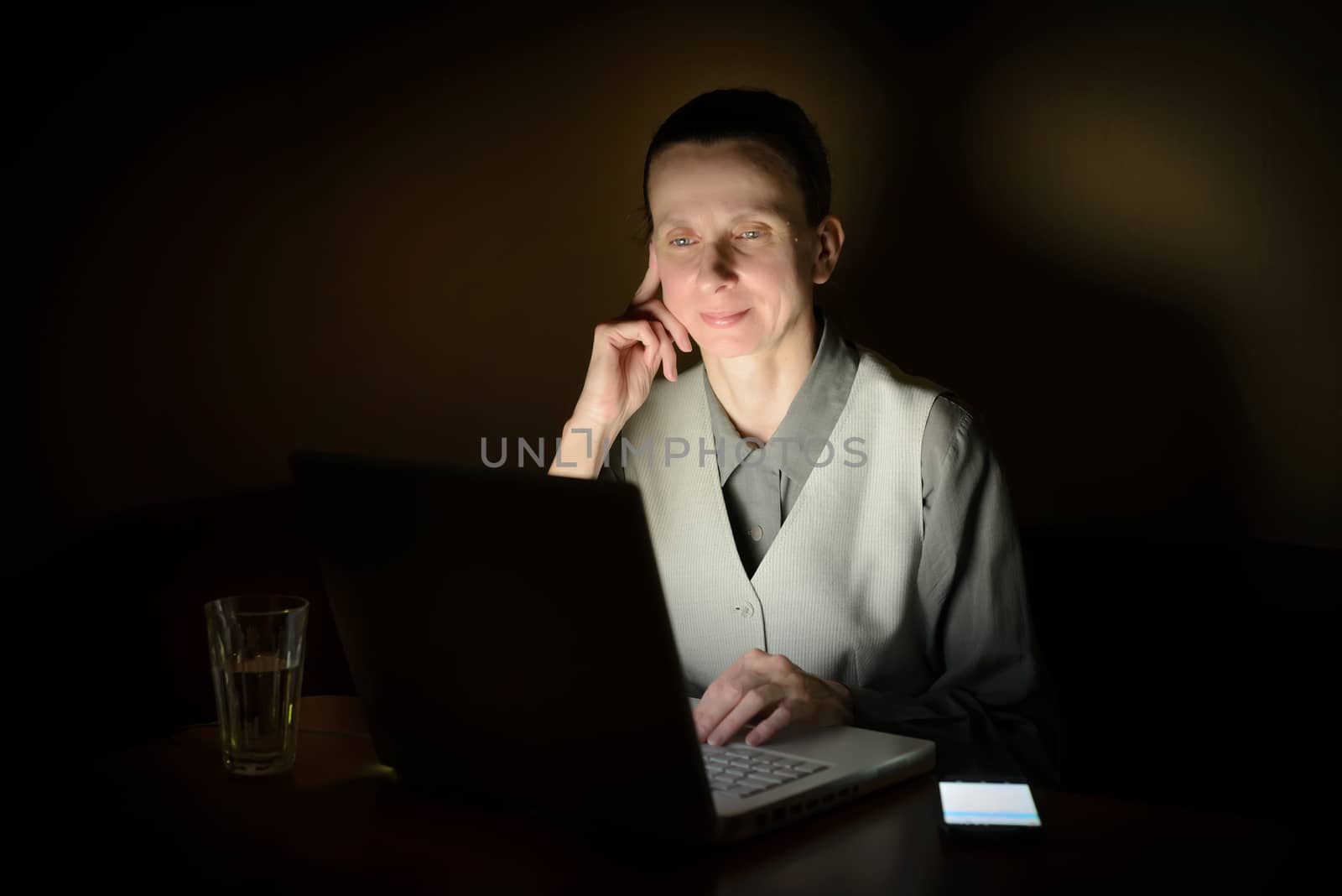 A business woman is using a computer in a dark room. Her face is illuminated by the light from the lcd monitor.
