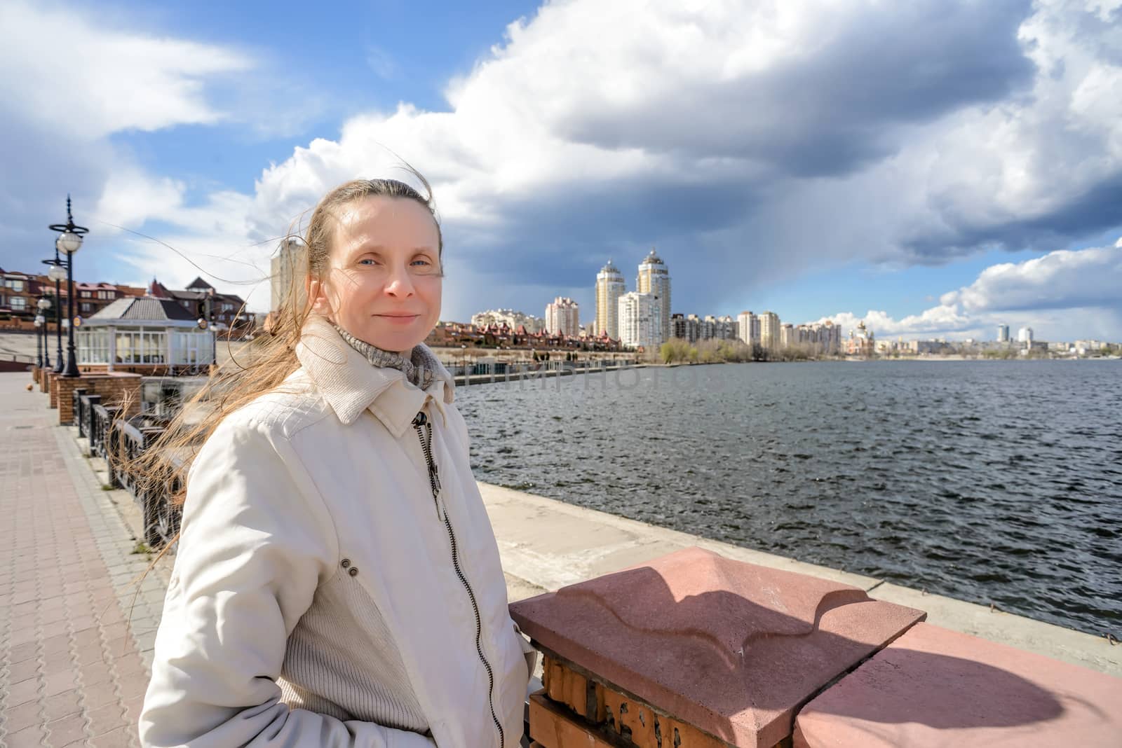 A smiling woman stands close to the river, her hair is moved by the wind under a dramatic stormy spring sky