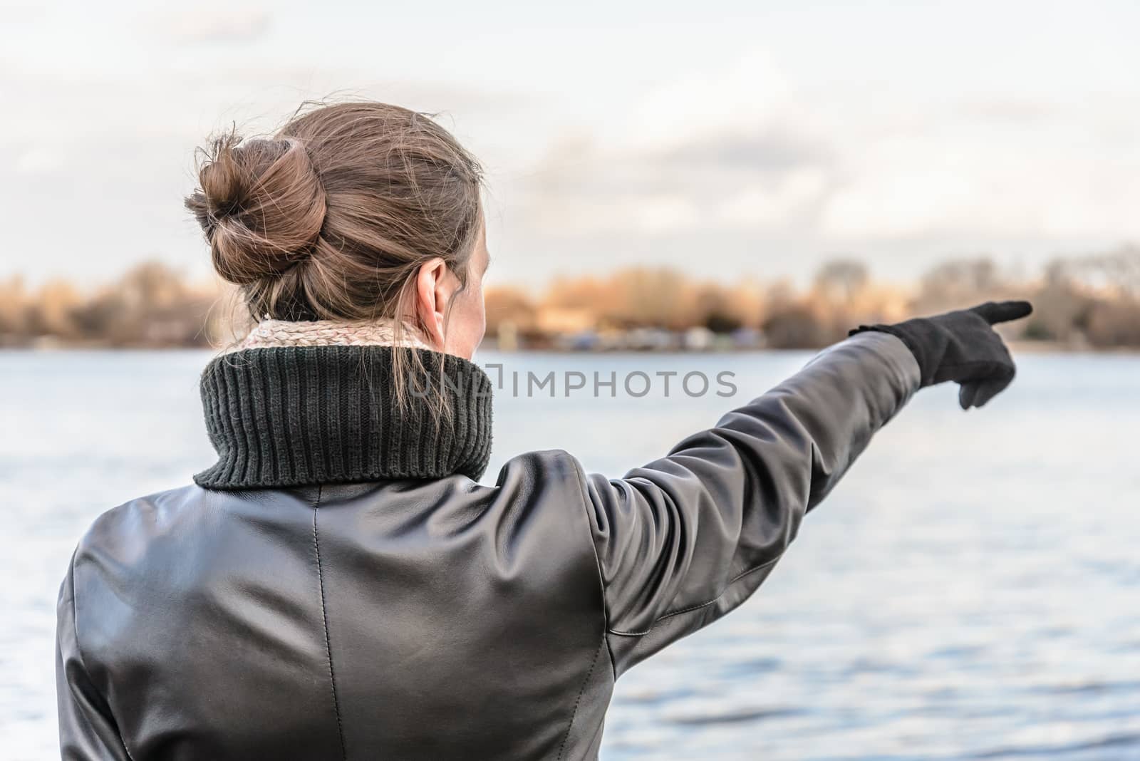 An adult with a chignon and wearing a black leather coat stays close to the river and point her finger to indicate something interesting on the opposite bank