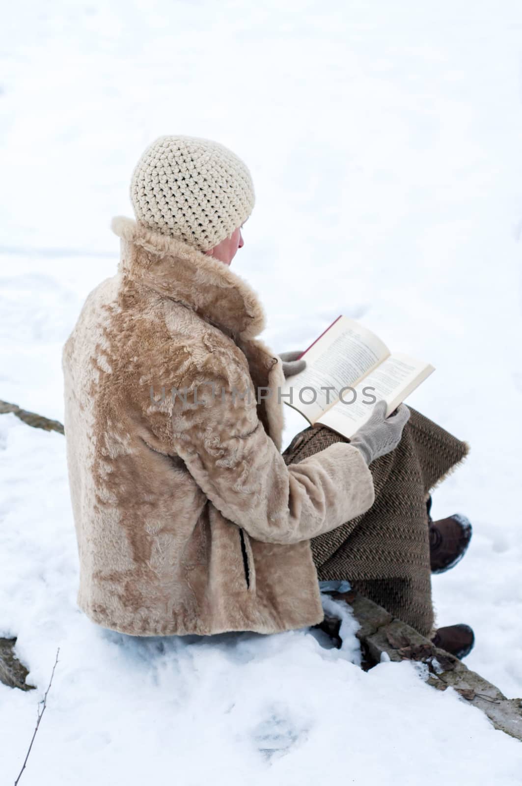 An adult woman with a fur coat and a woolen cap is sitting on a stone bench and reading a book in winter, in  a park covered by snow