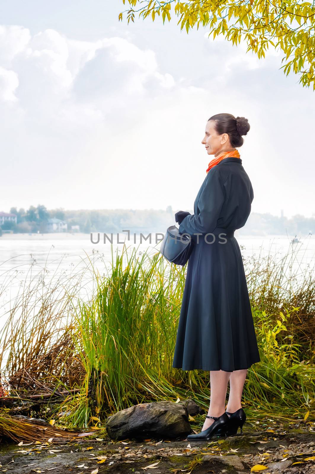 Elegant senior business woman with a bag and an orange scarf, walking along the river, under the trees in autumn