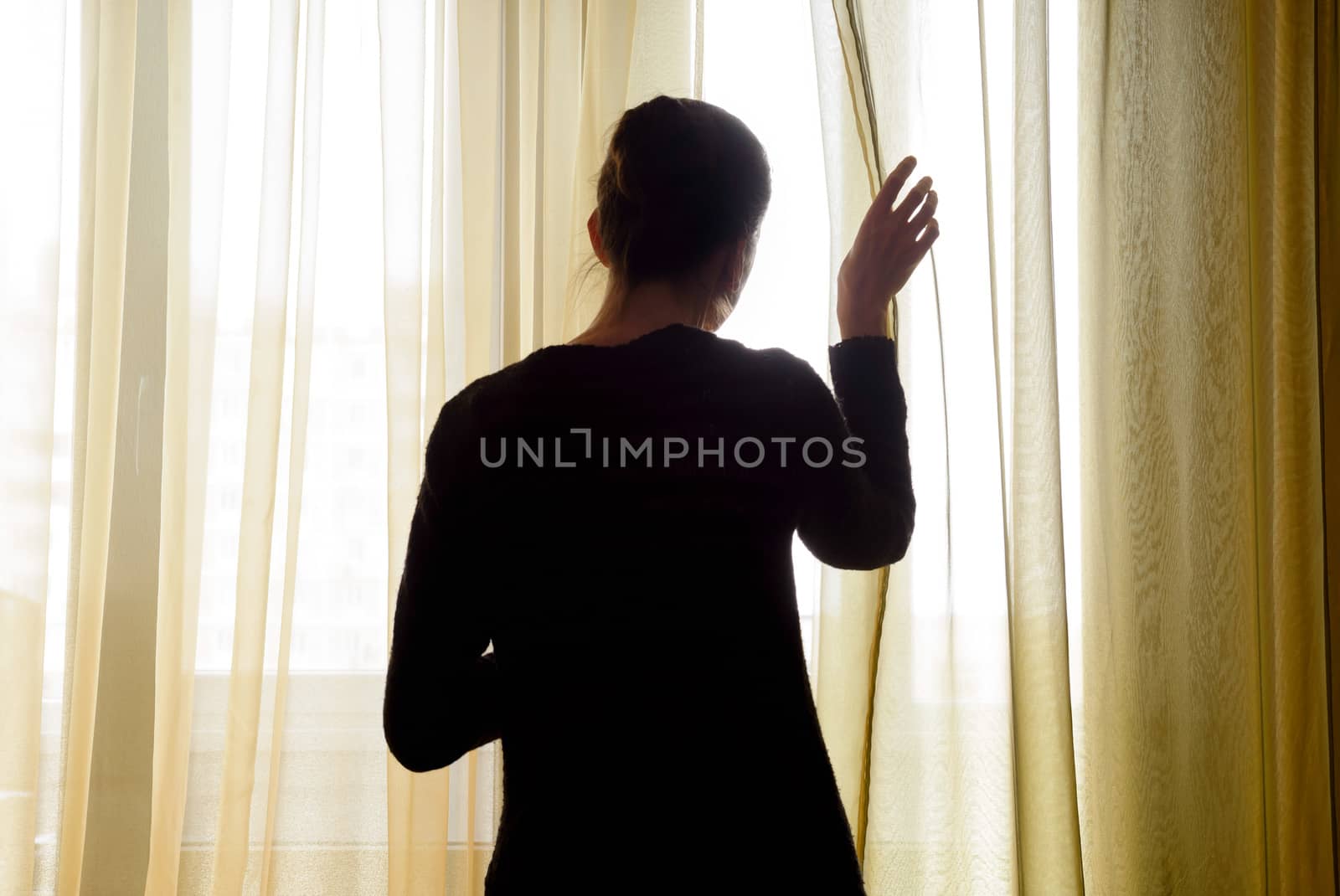 A woman in black moves the curtain aside to look out trough the window