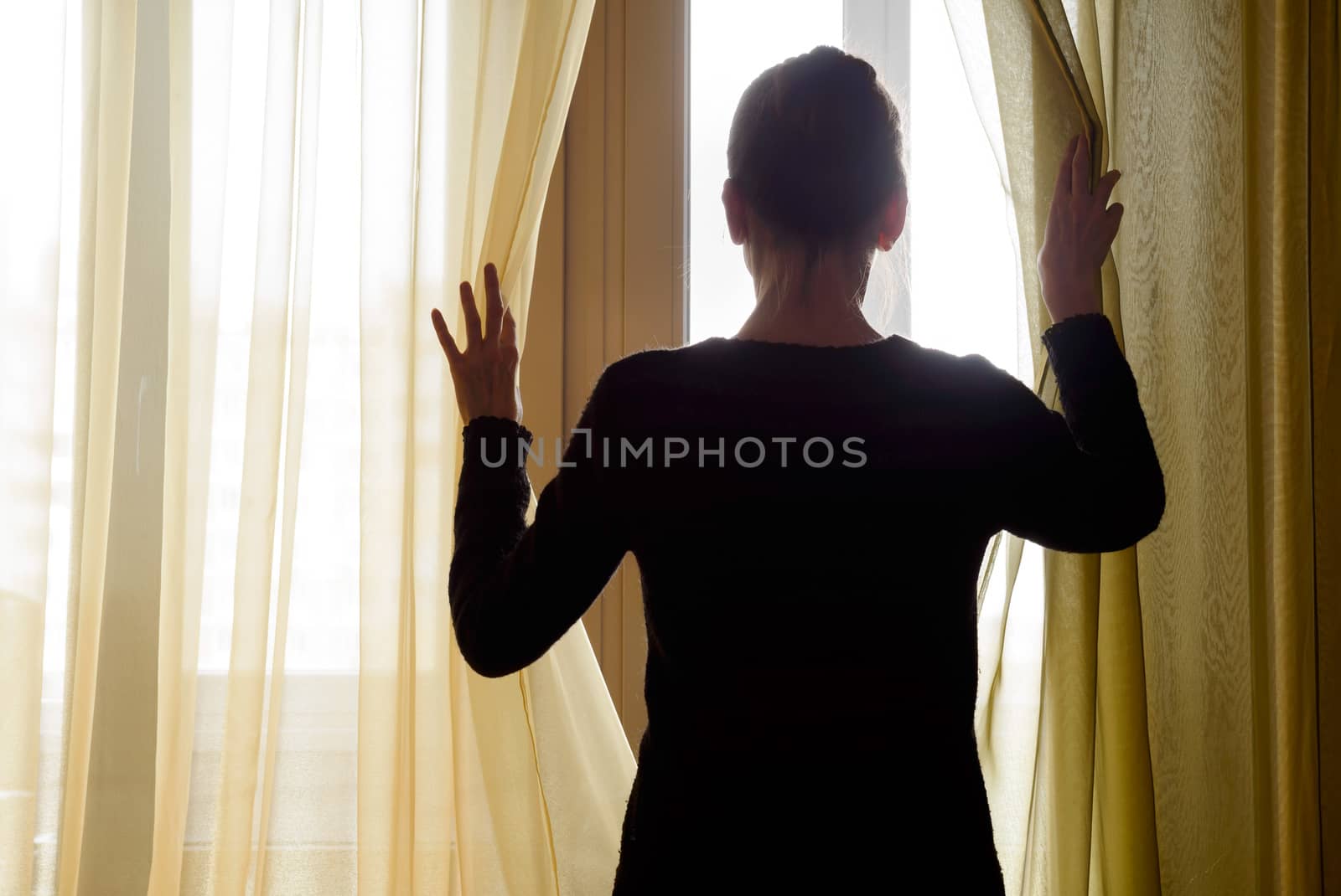 A woman in black moves the curtain aside to look out trough the window