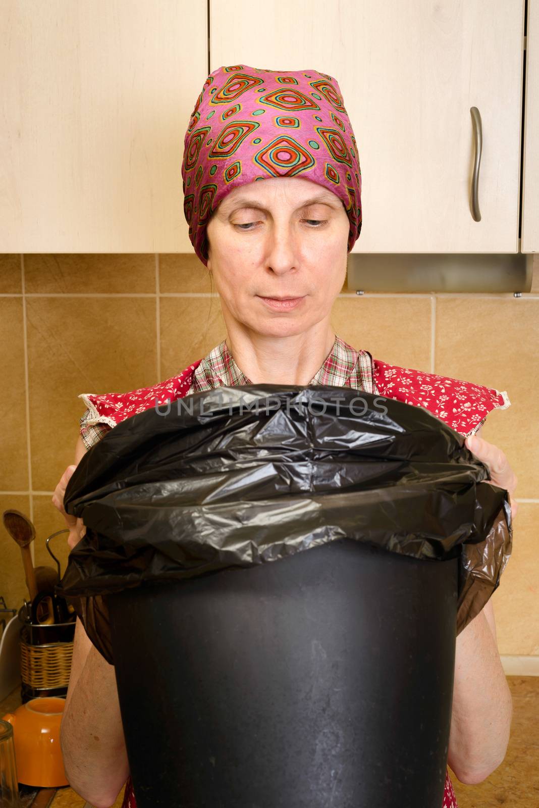 A woman, with a scarf on the head and a red apron, is looking inside a black trash can with a garbage bag, in the kitchen. She is very disturbed by the bad smell