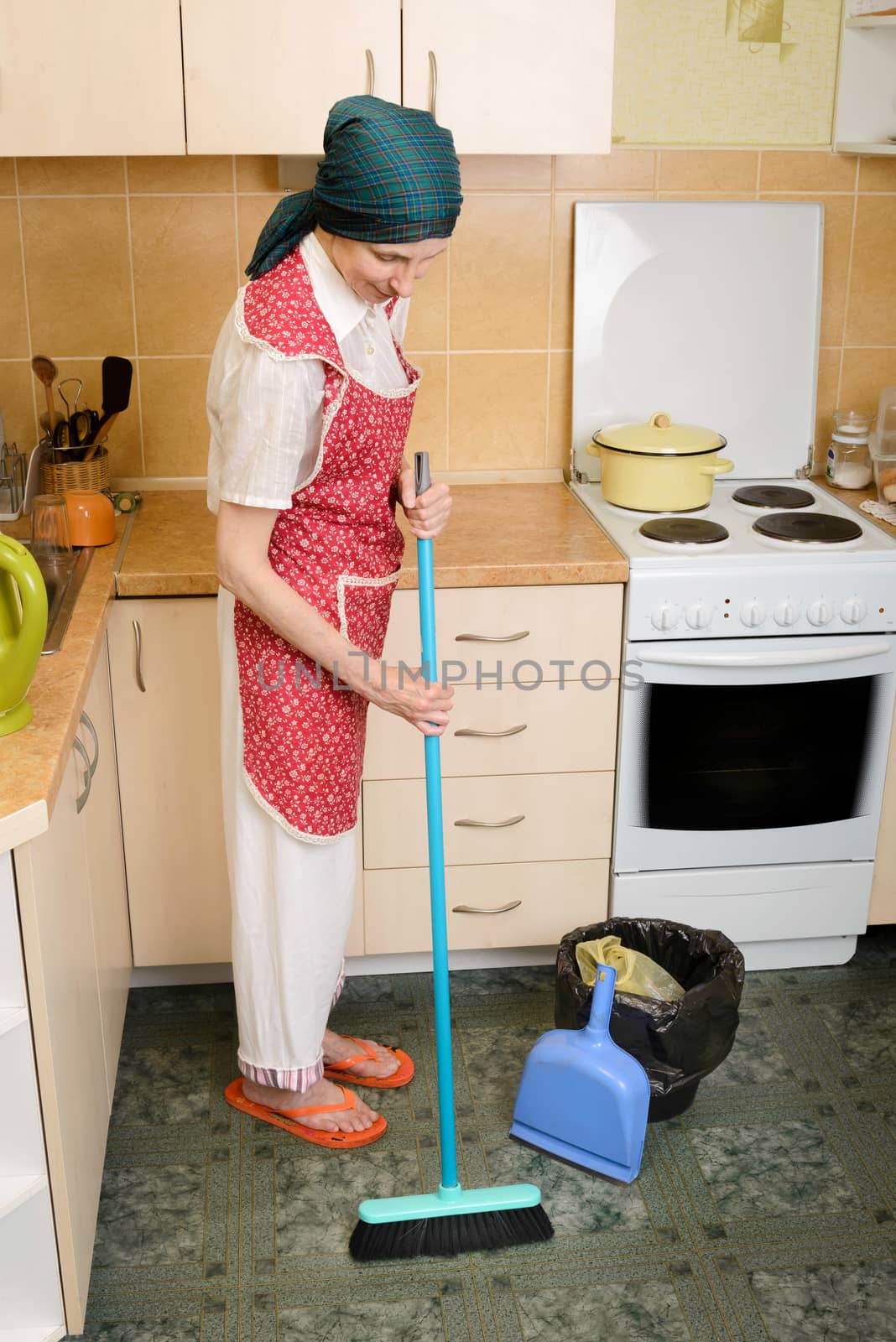 An  adult woman, a housewife or a maid, wearing a red apron and a green scarf on her head is sweeping the kitchen with a broom