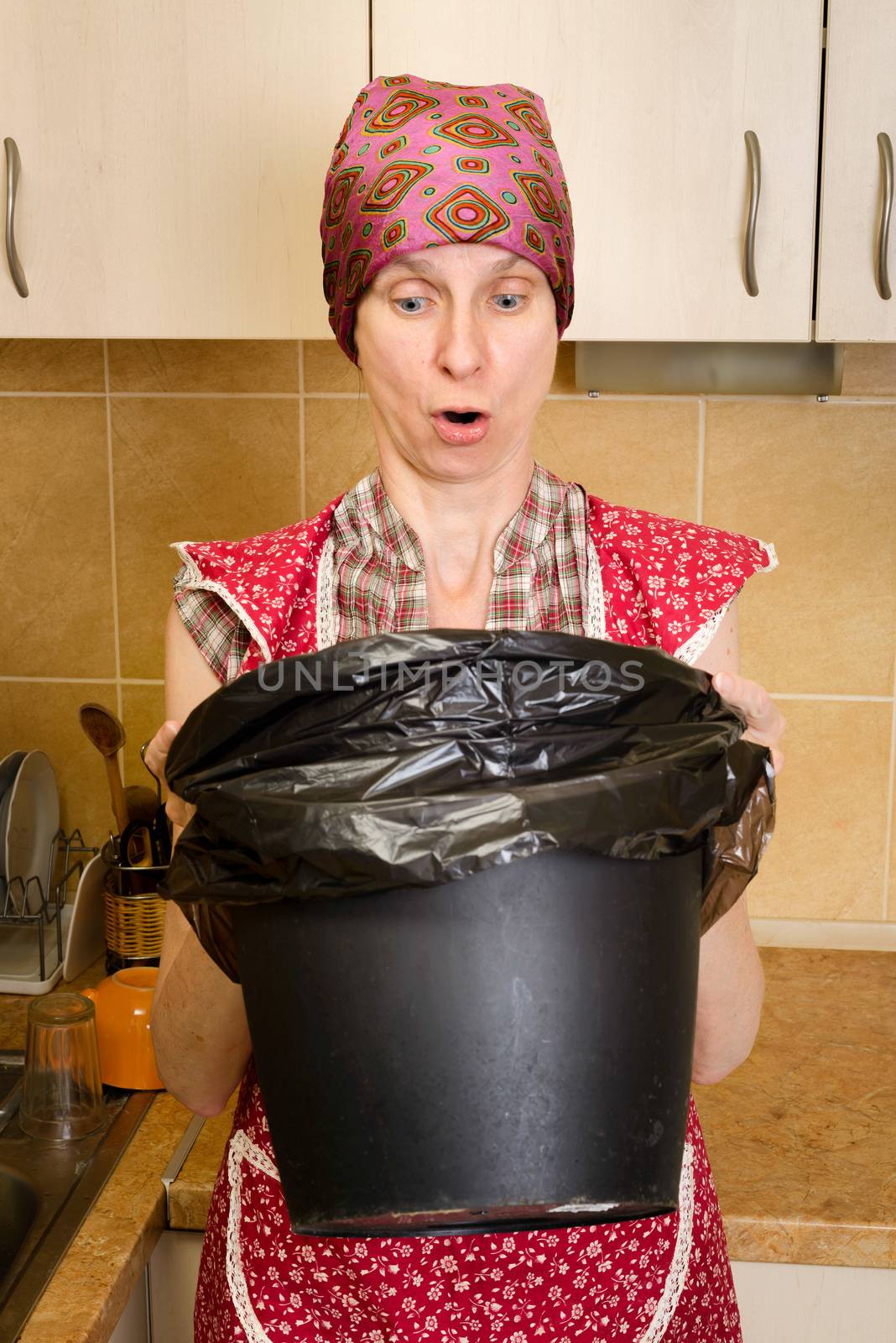 A woman, with a scarf on the head and a red apron, is looking inside a black trash can with a garbage bag, in the kitchen. She is very disturbed by the bad smell