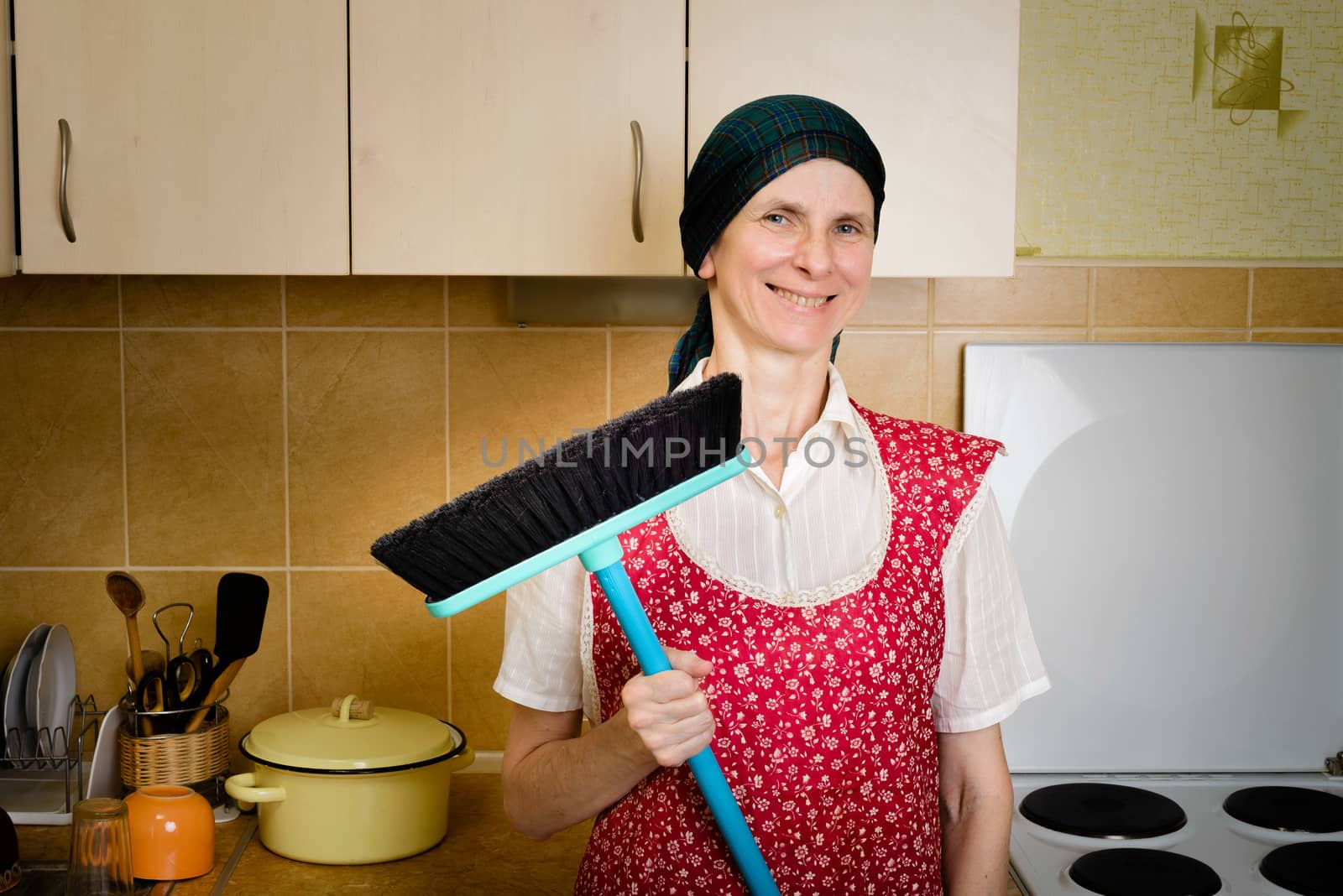 A happy adult woman portrait, a housewife or a maid, wearing a red apron and a green scarf on her head is resting after she has swept the kitchen with a broom