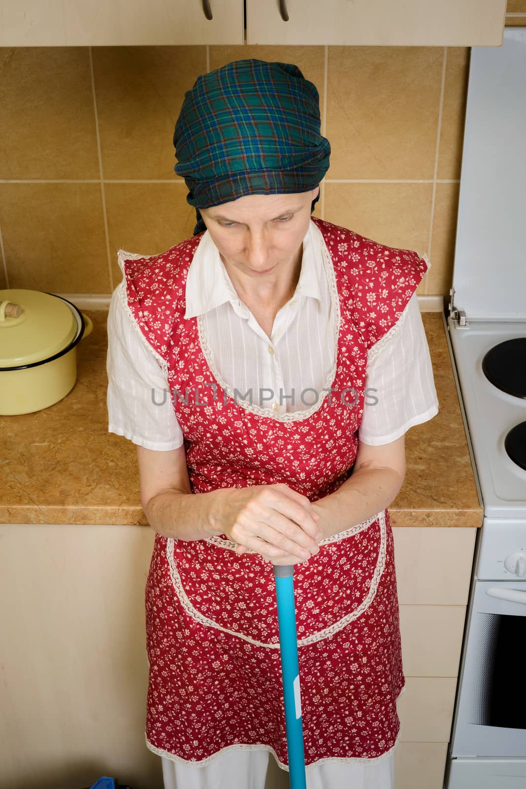 A depressed  adult woman, a housewife or a maid, wearing a red apron and a green scarf on her head is resting after she has swept the kitchen with a broom