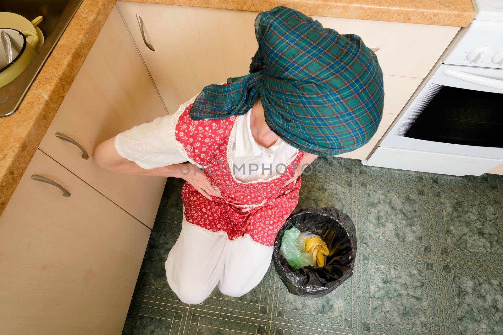 A woman, with a scarf on the head and a red apron, is looking inside a black trash can with a garbage bag, in the kitchen. She is very disturbed by the bad smell