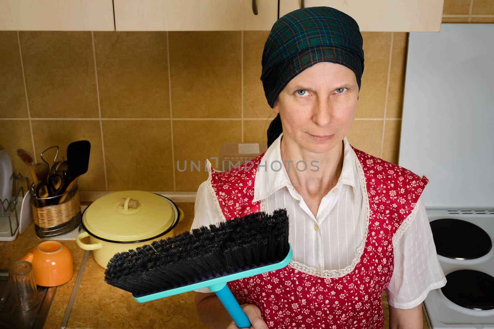 An angry and tired adult woman portrait, a housewife, a maid or maybe a witch, wearing a red apron and a green scarf on her head. She does not want to sweep the kitchen with her broom.