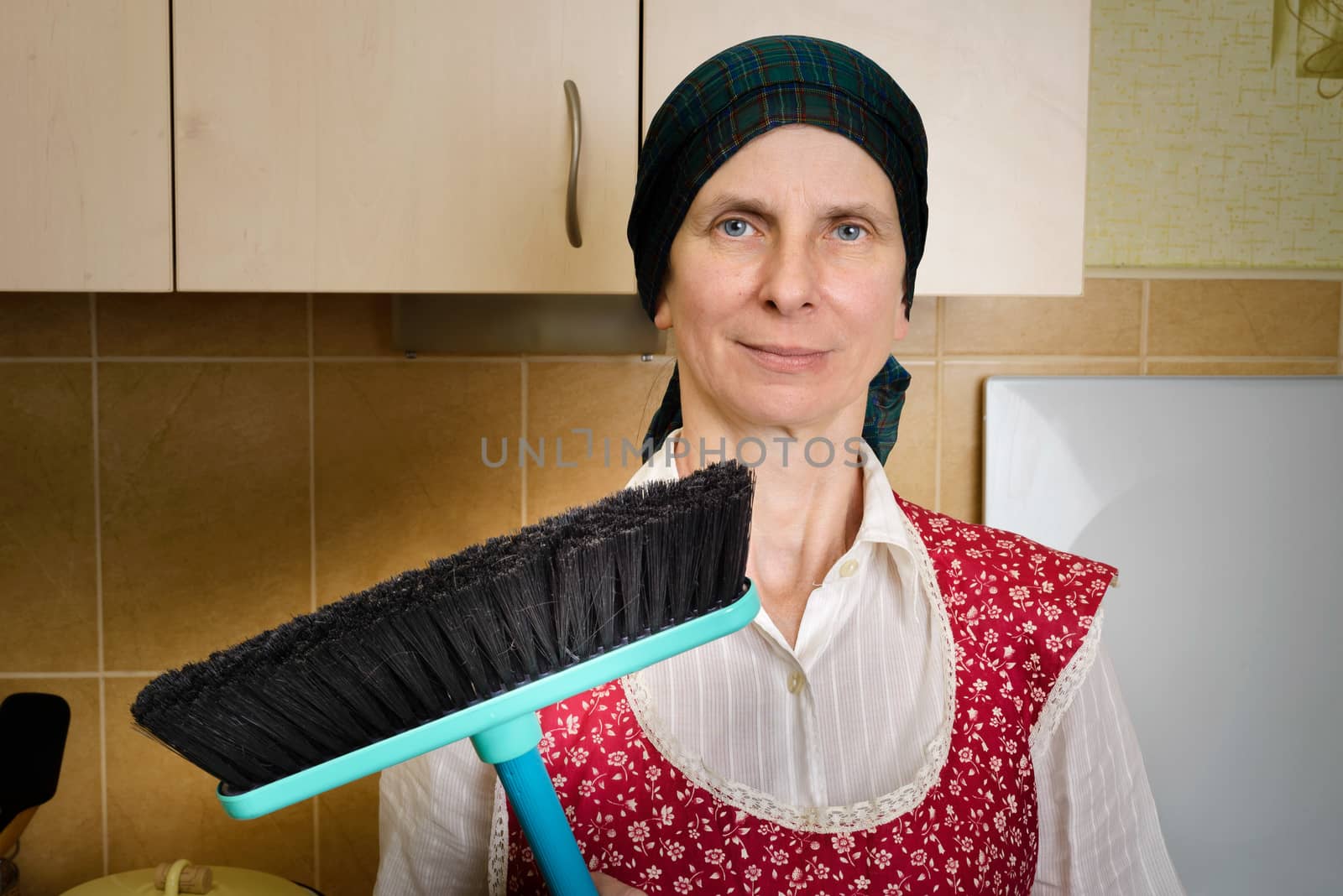 A happy adult woman portrait, a housewife or a maid, wearing a red apron and a green scarf on her head is resting after she has swept the kitchen with a broom