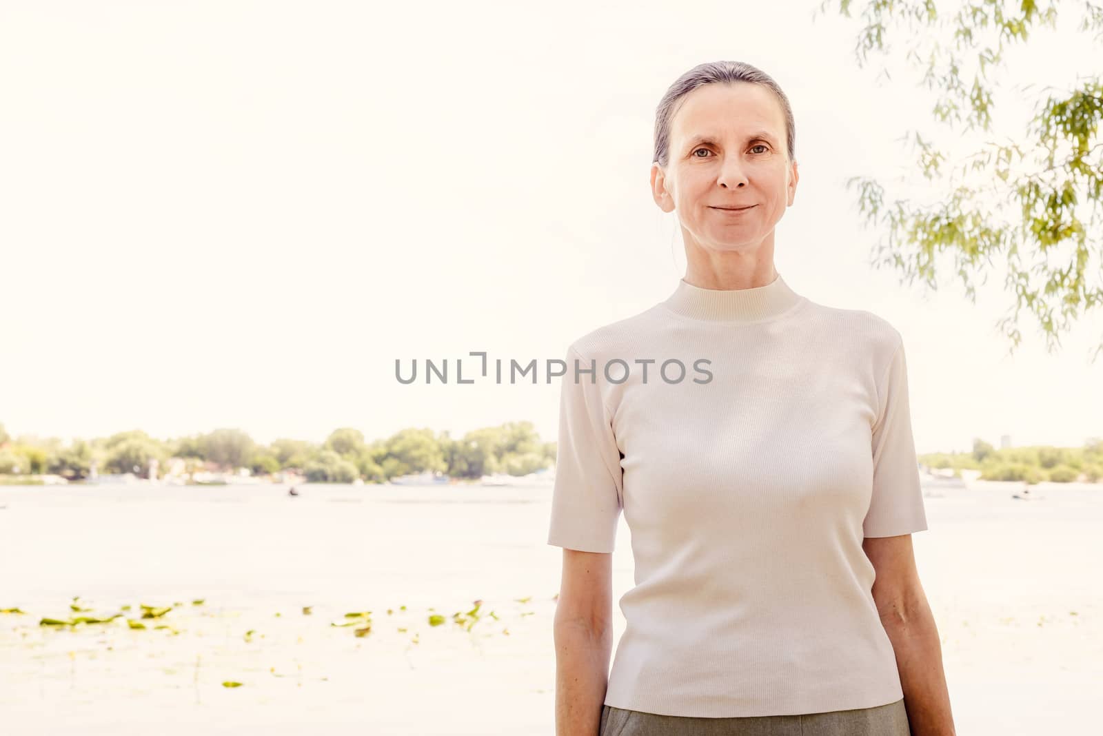 A warm portrait of a nice smiling senior woman wearing a white jumper close to the river in summer