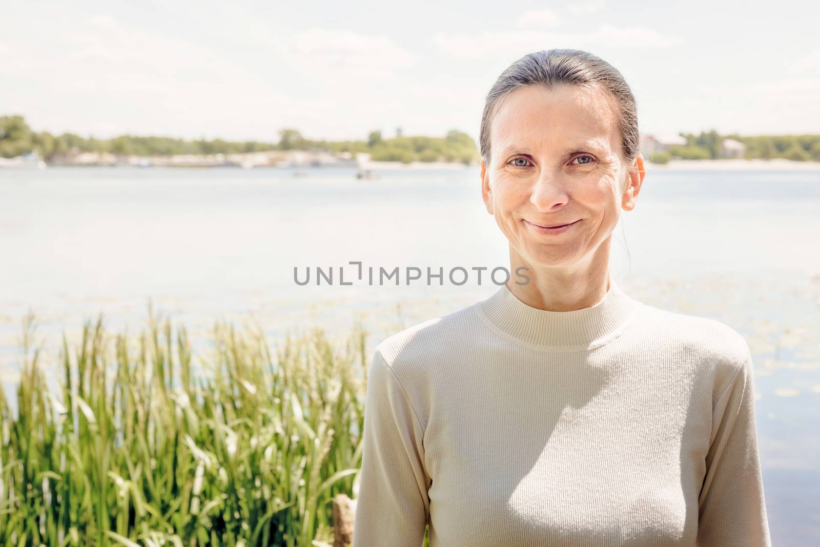 A warm portrait of a nice smiling senior woman wearing a white jumper close to the river in summer