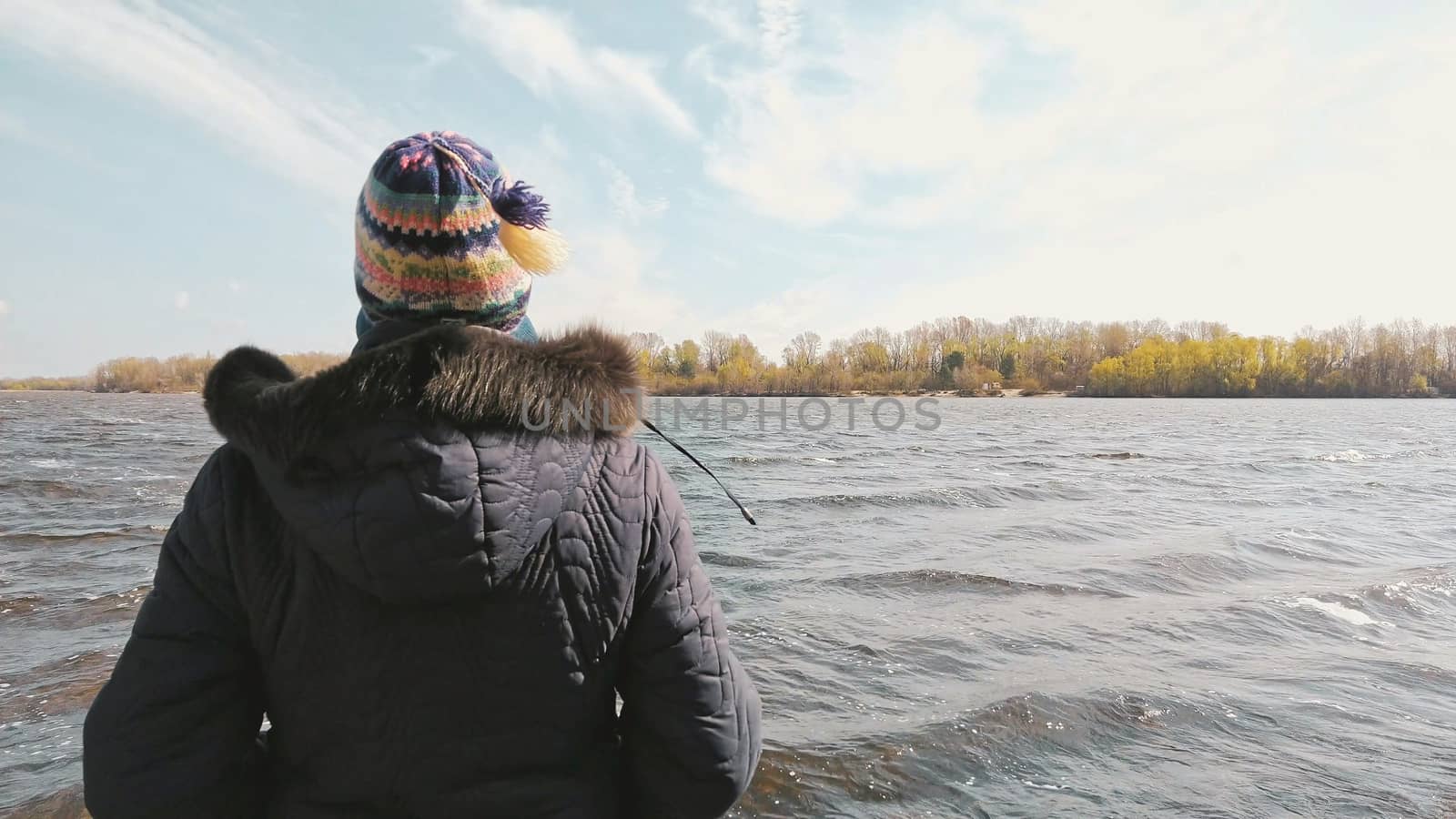 A woman with a woolen cap and a warm coat is looking at the Dnieper river, in Kiev, Ukraine, during a cold and sunny spring morning. The wind and the stream move the water, creating waves