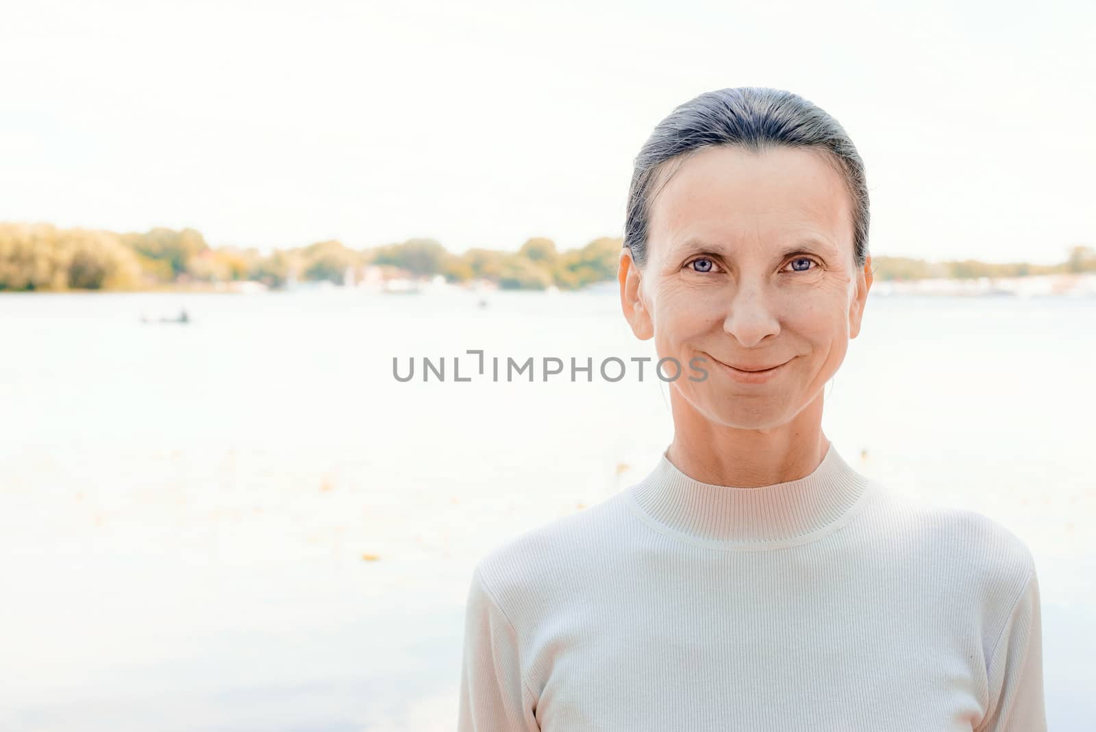 A warm portrait of a nice smiling senior woman wearing a white jumper close to the river in summer