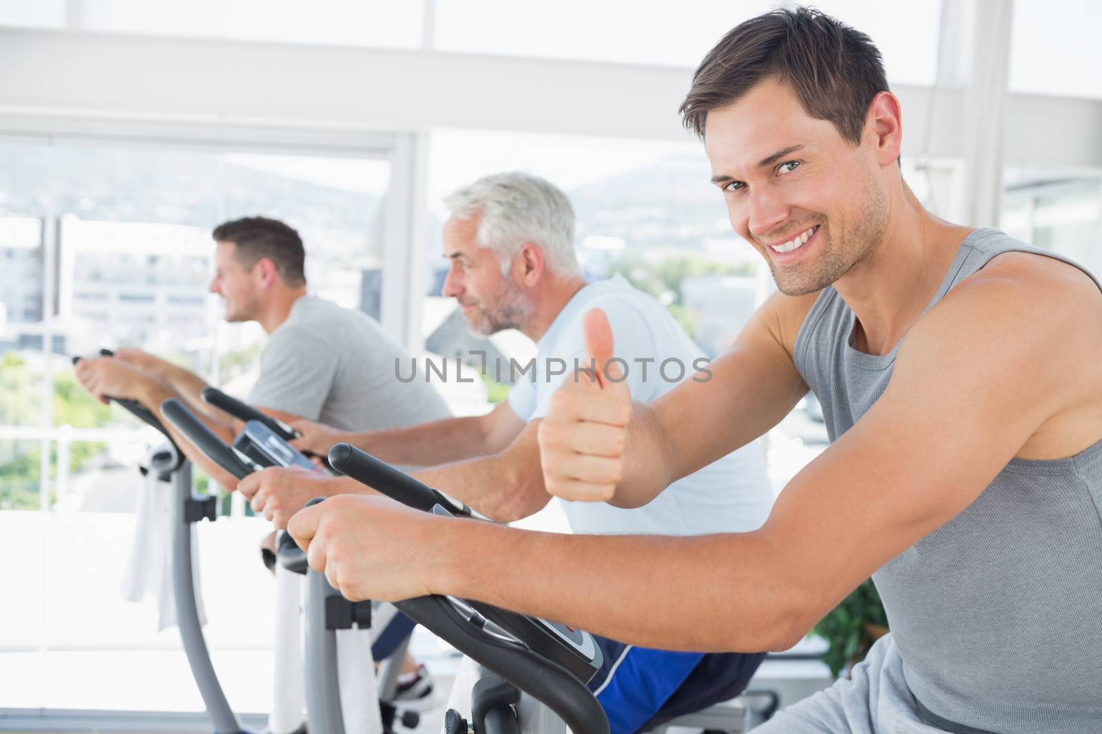 Portrait of happy man on exercise bike gesturing thumbs up at gym
