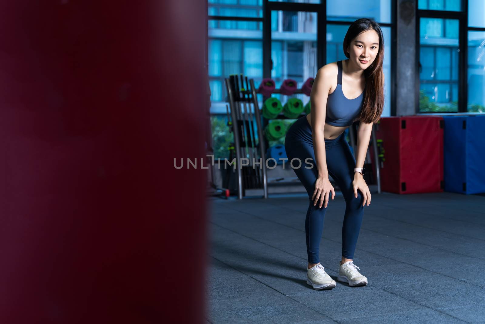 Asian Women rest during exercise. She's holding her hand at Knee with blur Racks Fitness Equipment background xhaustion and being alone at the gym sport and healthy concept