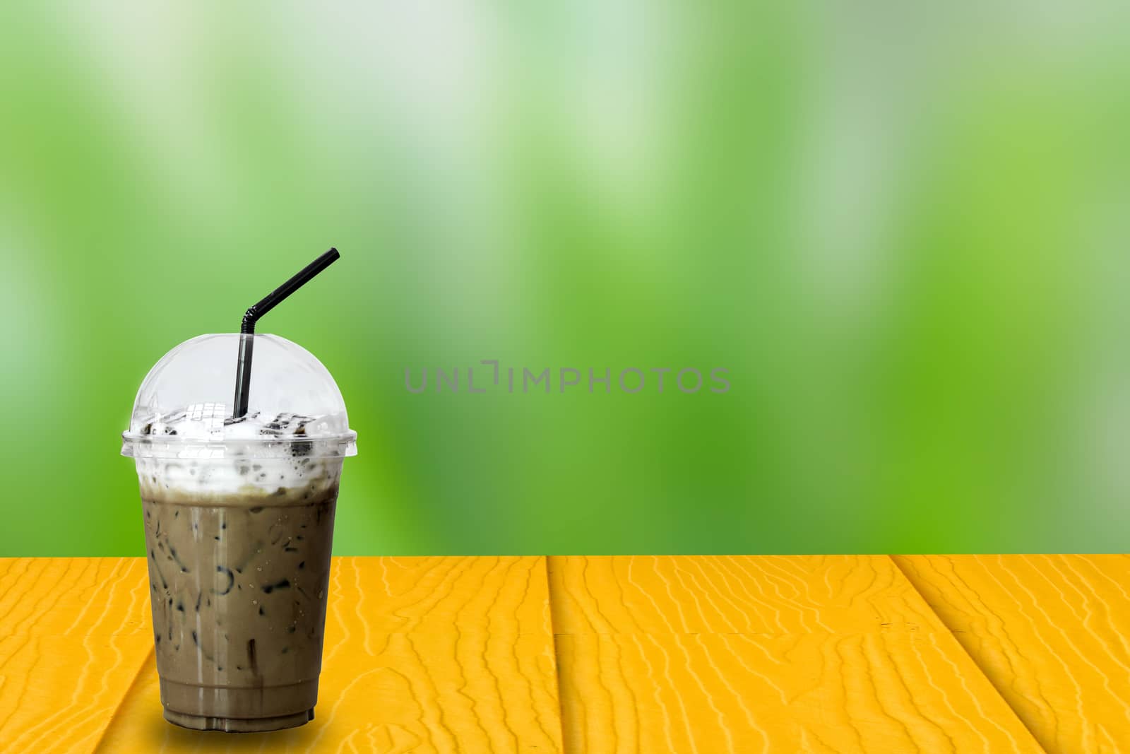  Ice Coffee in plastic take away cup with black straw on yellow wooden table on green nature blurry background, copy space for your text. Montage product display.