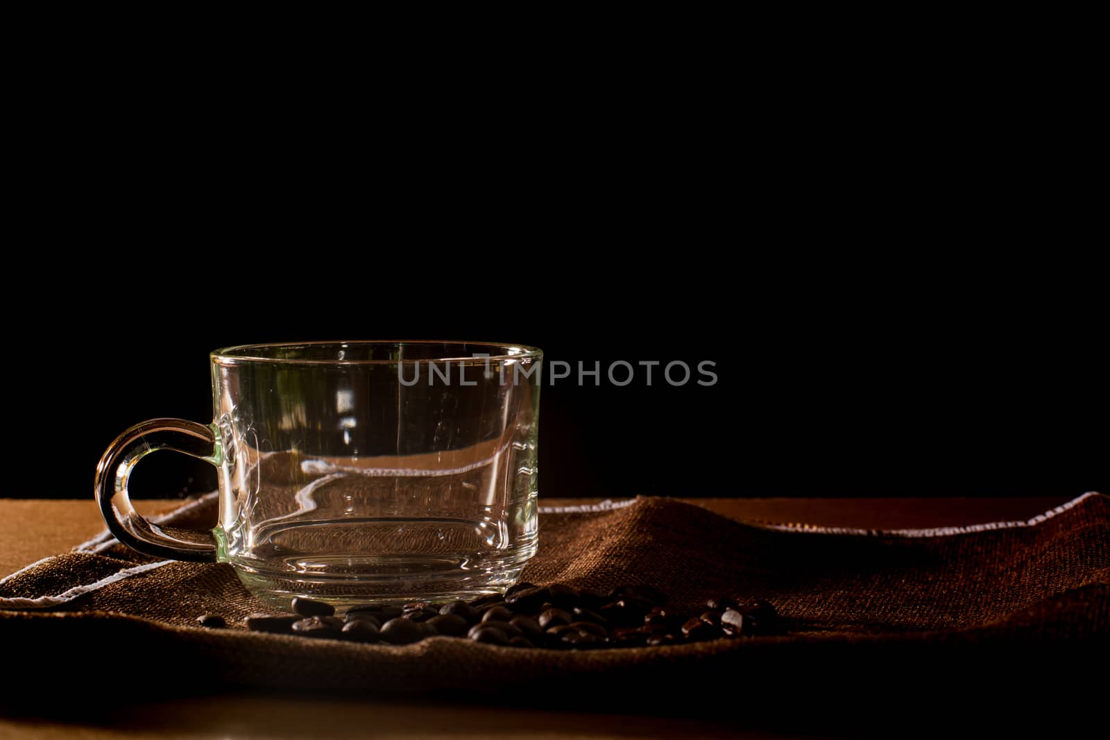 Empty cup and group of coffee beans on brown table cloth with black background with copy space for your text. Benefits of coffee concept. 