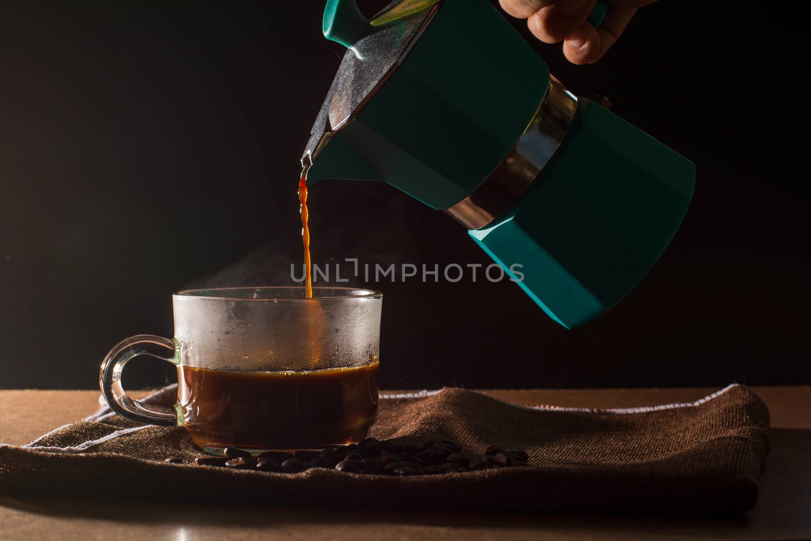 Pour hot of black coffee from green moka pot to clear coffee cup with smoke and coffee beans on brown table cloth and wooden table. Benefit of coffee concept.