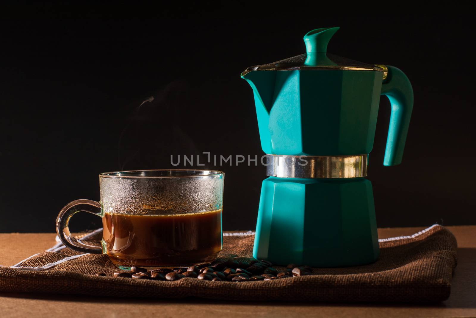 Close up hot of black coffee and green moka pot and coffee beans on brown table cloth and wooden table. Benefit of coffee concept. 