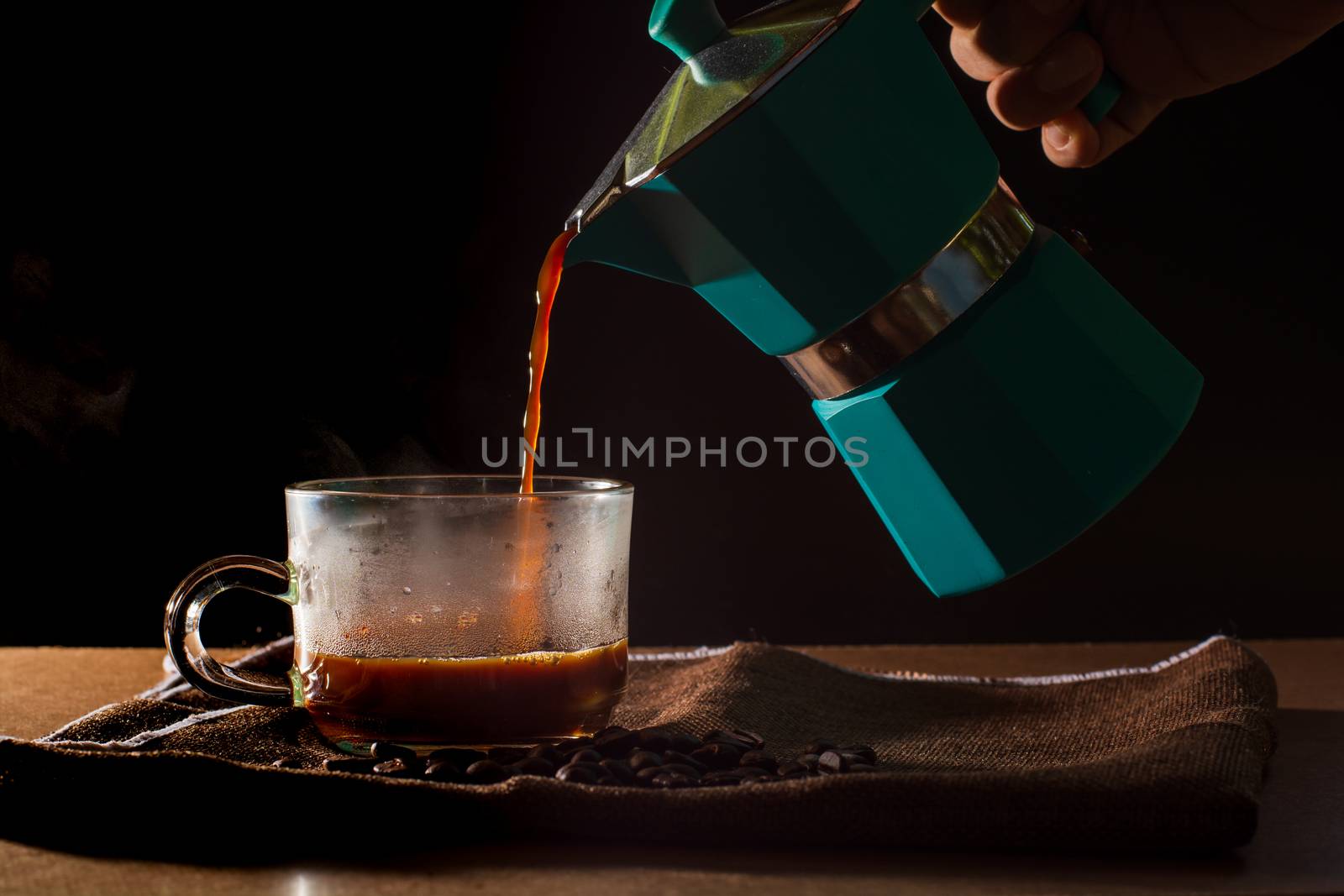 Pour hot of black coffee from green moka pot to clear coffee cup with smoke and coffee beans on brown table cloth and wooden table. Benefit of coffee concept.