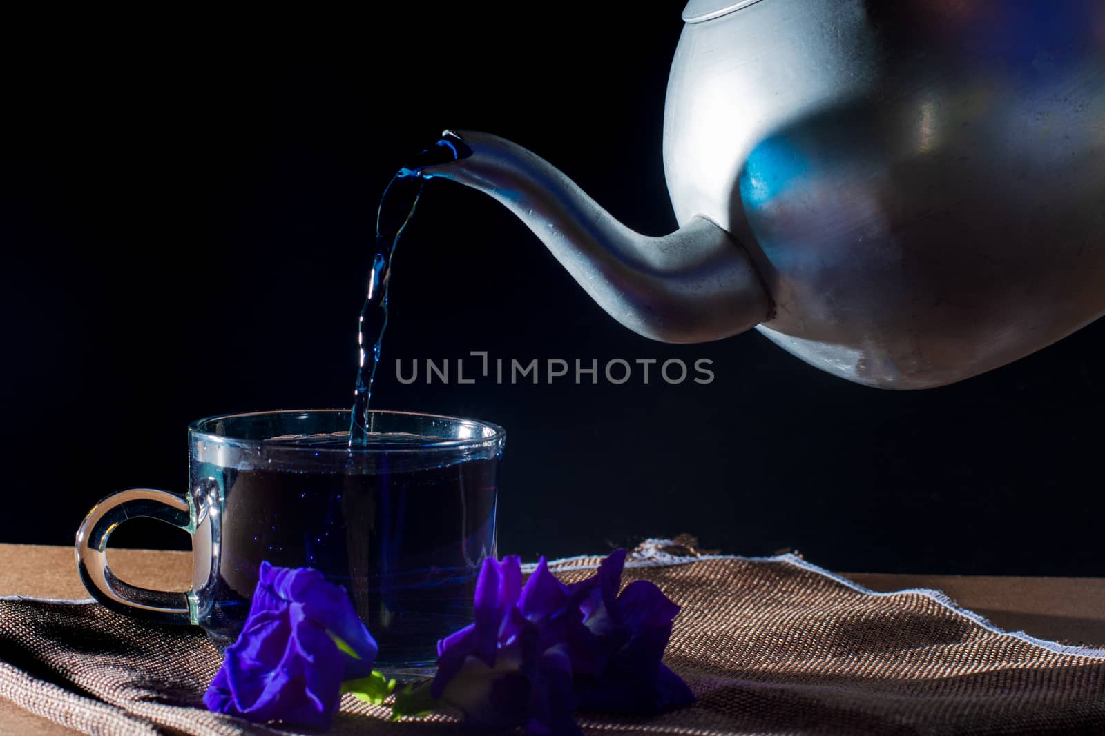 Pour Butterfly pea tea by metal pot to cup with violet flower on brown table cloth on black background. Healthy beverage for drink. Herbs and medical concept. 

