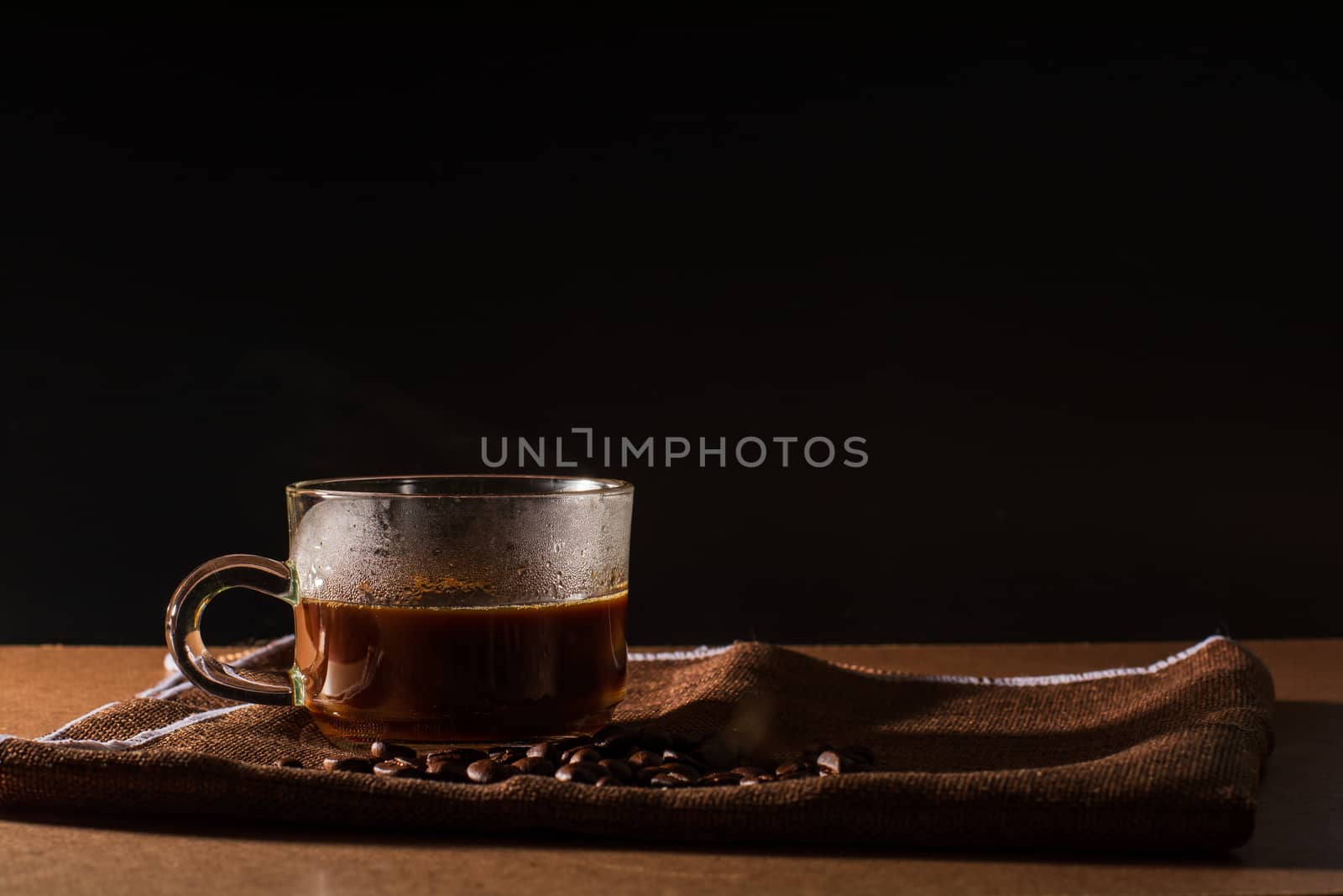 Cup of hot coffee with smoke and group of coffee beans on brown table cloth with black background with copy space for your text. Benefits of coffee concept.