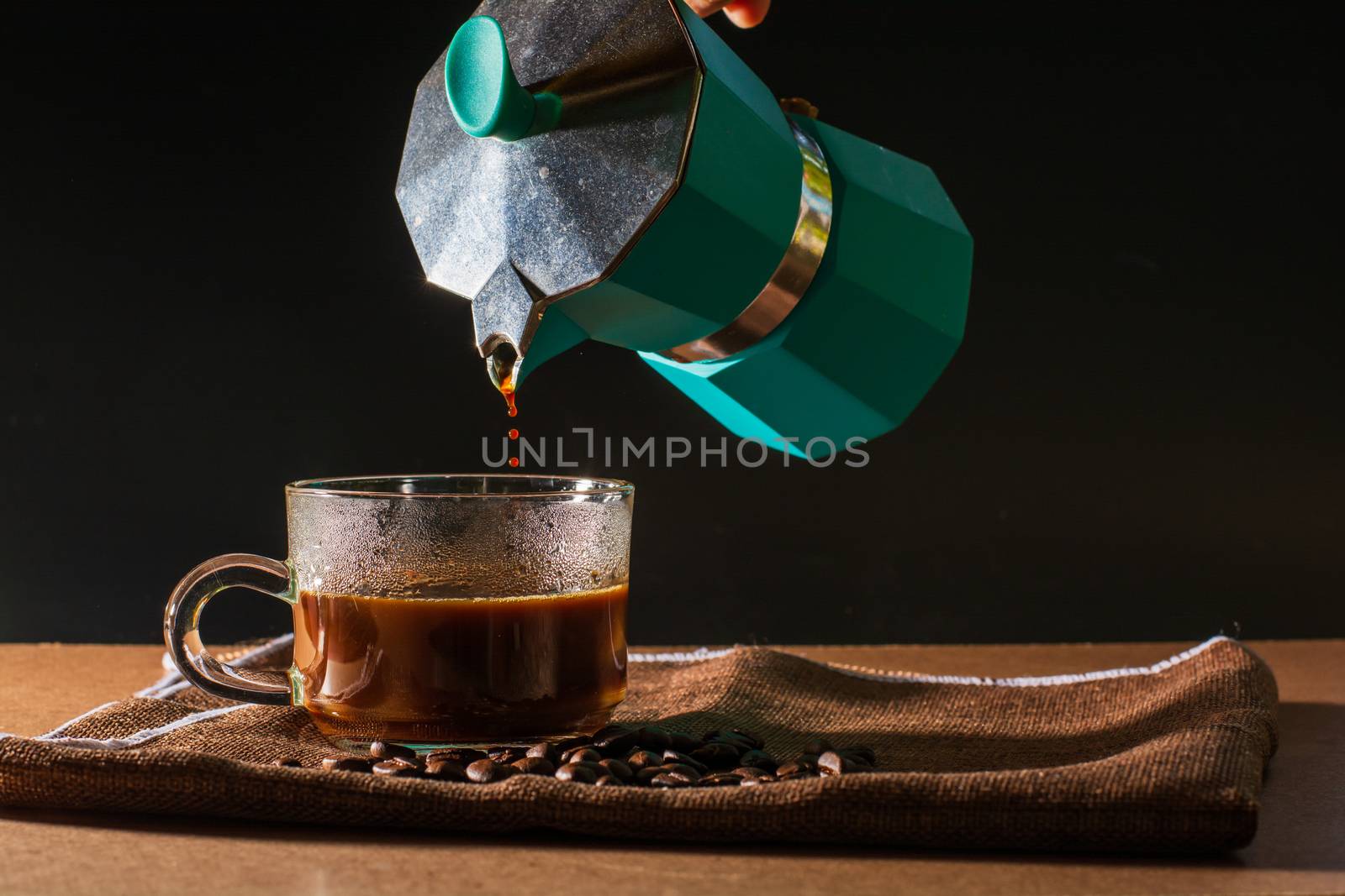 Pour hot of black coffee from green moka pot to clear coffee cup with smoke and coffee beans on brown table cloth and wooden table. Benefit of coffee concept.