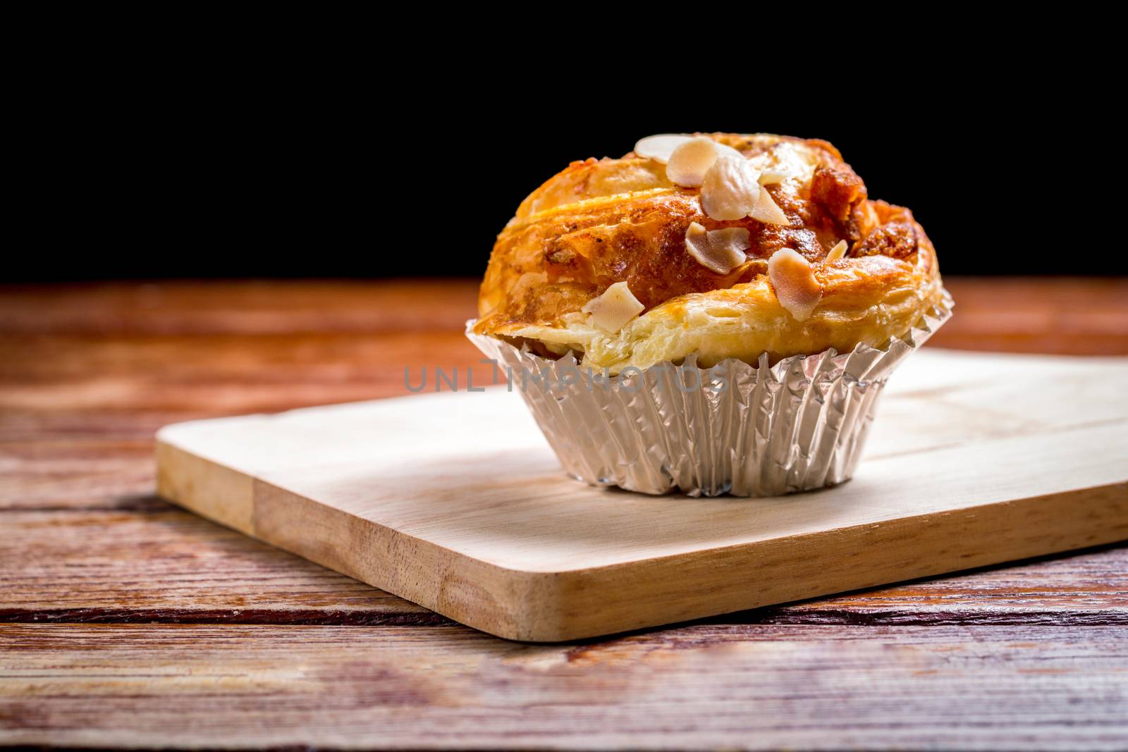 Delicious homemade almond danish bread in foil cup on a wooden chopping board and table in the home kitchen on black background.