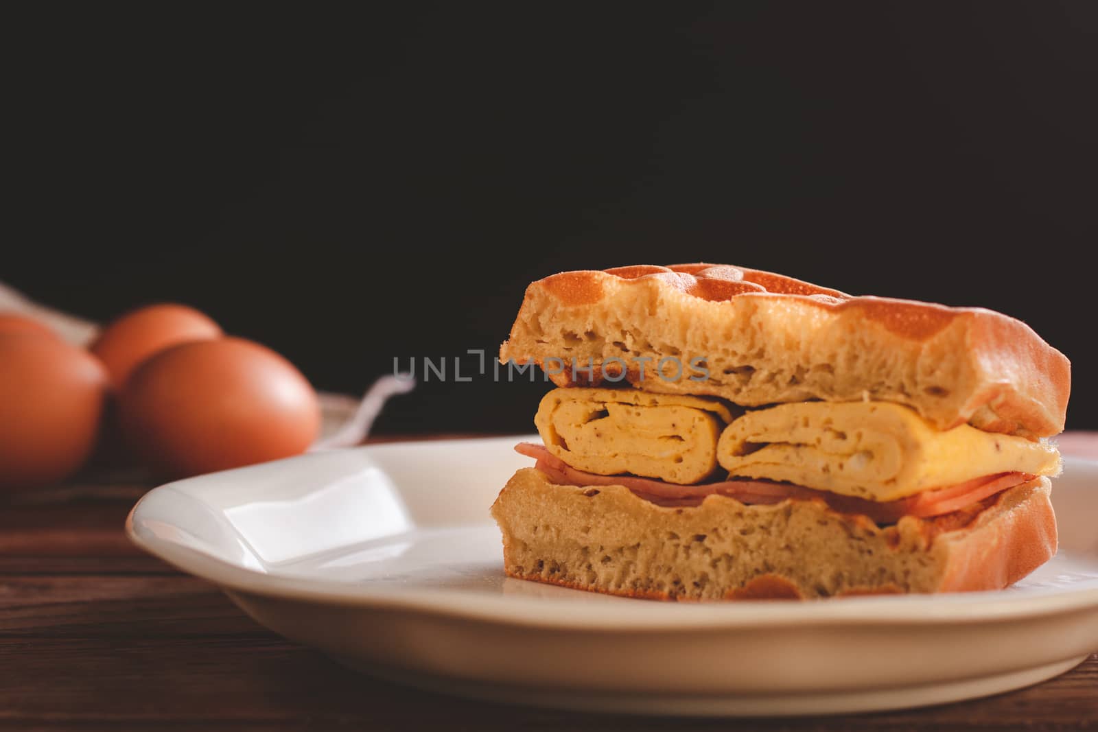 Waffle sandwich with egg and chicken ham in white dish on wooden table and black background. Homemade food concept.