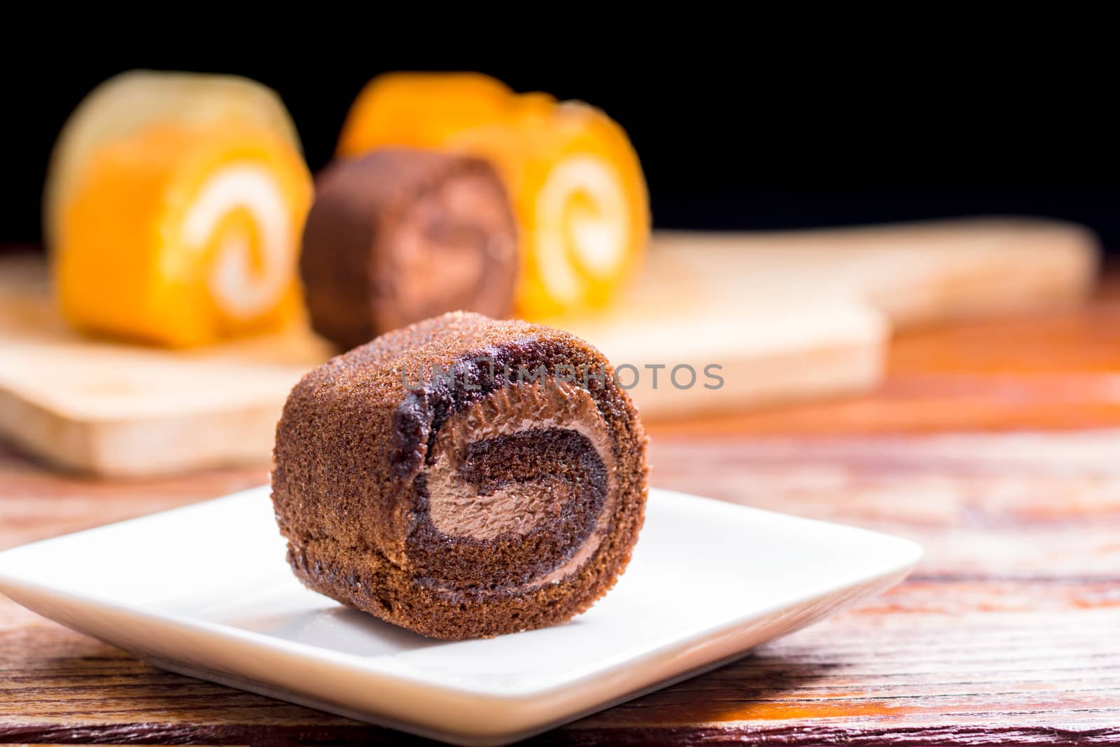 Close up delicious chocolate roll cake with whipping cream in a square white plate on a wooden table at the home kitchen with a black background and copy space. Homemade bakery concept.